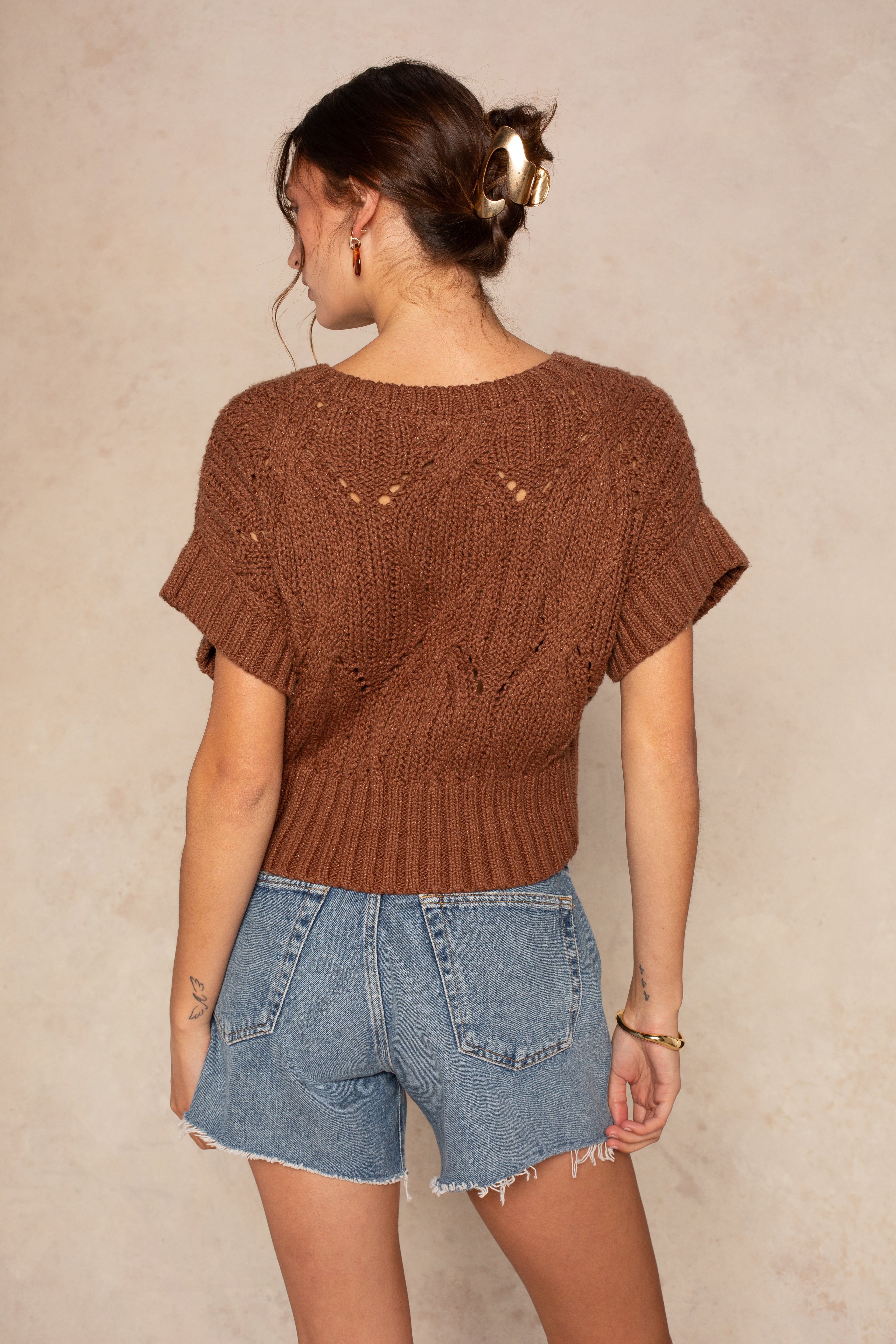 A woman with brown hair in a bun wears the Isla Cali Crochet chunky knit sweater and high-waisted, light-wash denim shorts, standing with her back to the camera against a beige background.