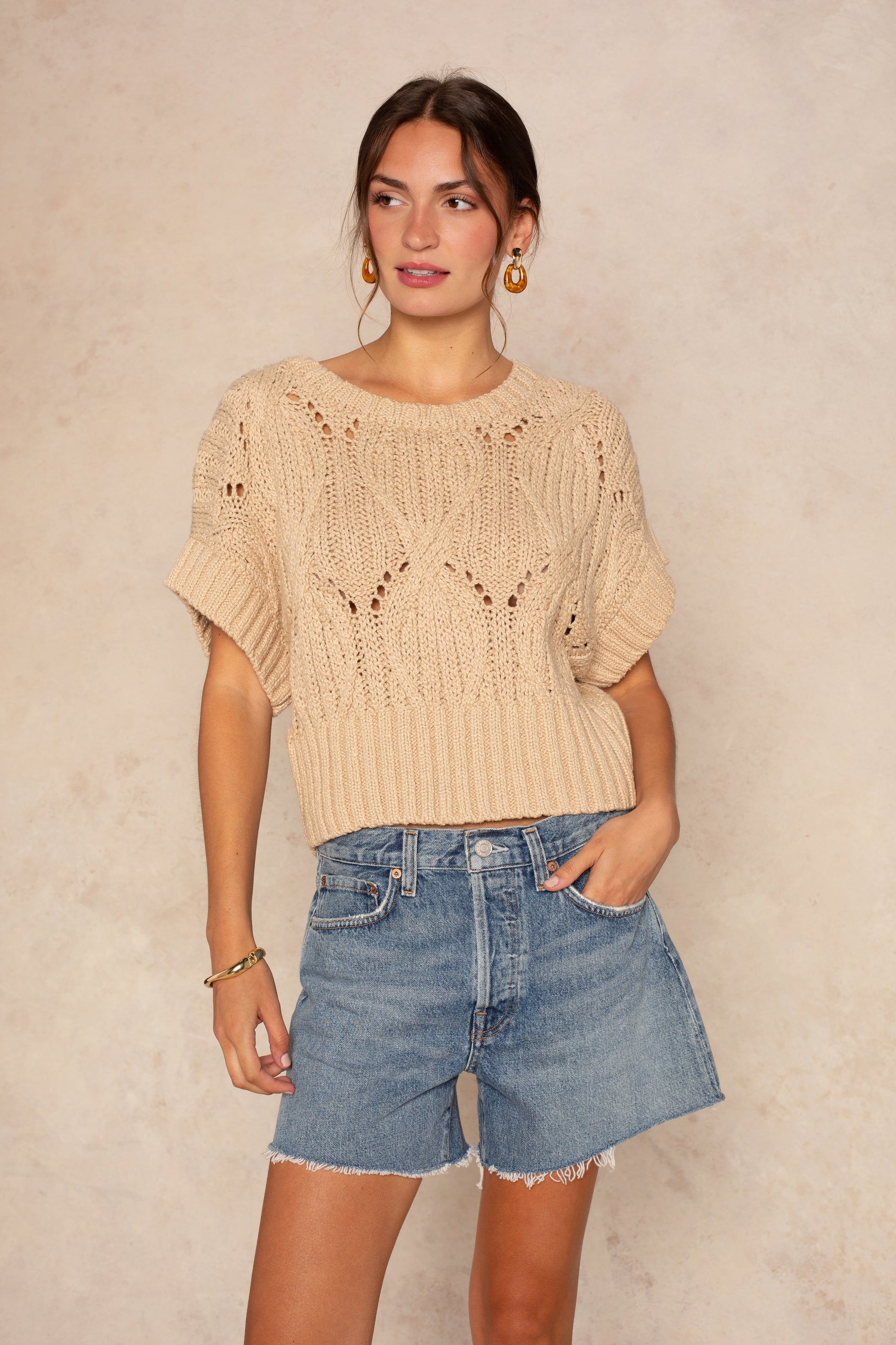 A woman wears the Isla "Cali Crochet" sweater in taupe with blue denim shorts, gold hoop earrings, and one hand in her pocket, standing against a neutral background.