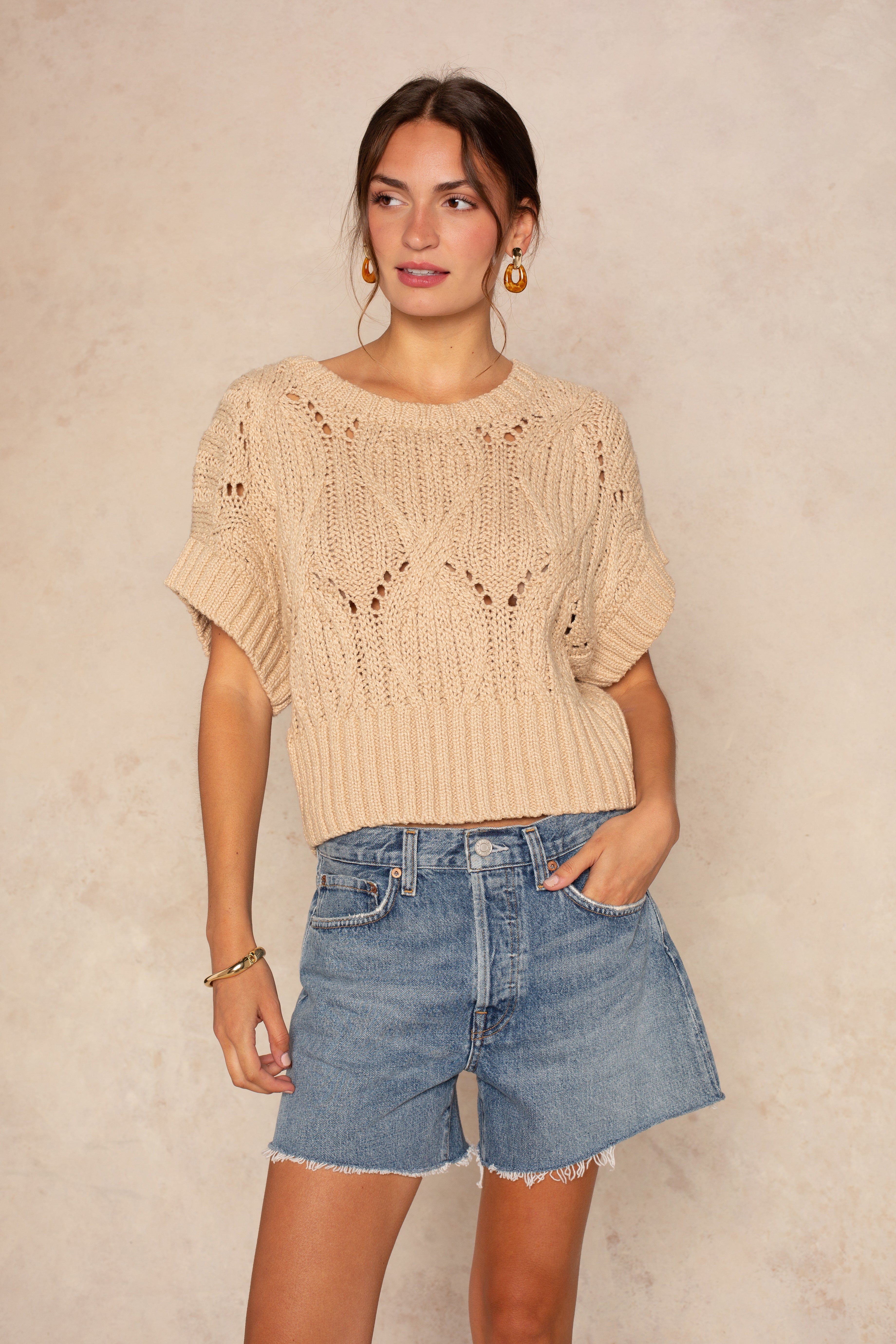 A woman wears the Isla "Cali Crochet" sweater in taupe with blue denim shorts, gold hoop earrings, and one hand in her pocket, standing against a neutral background.