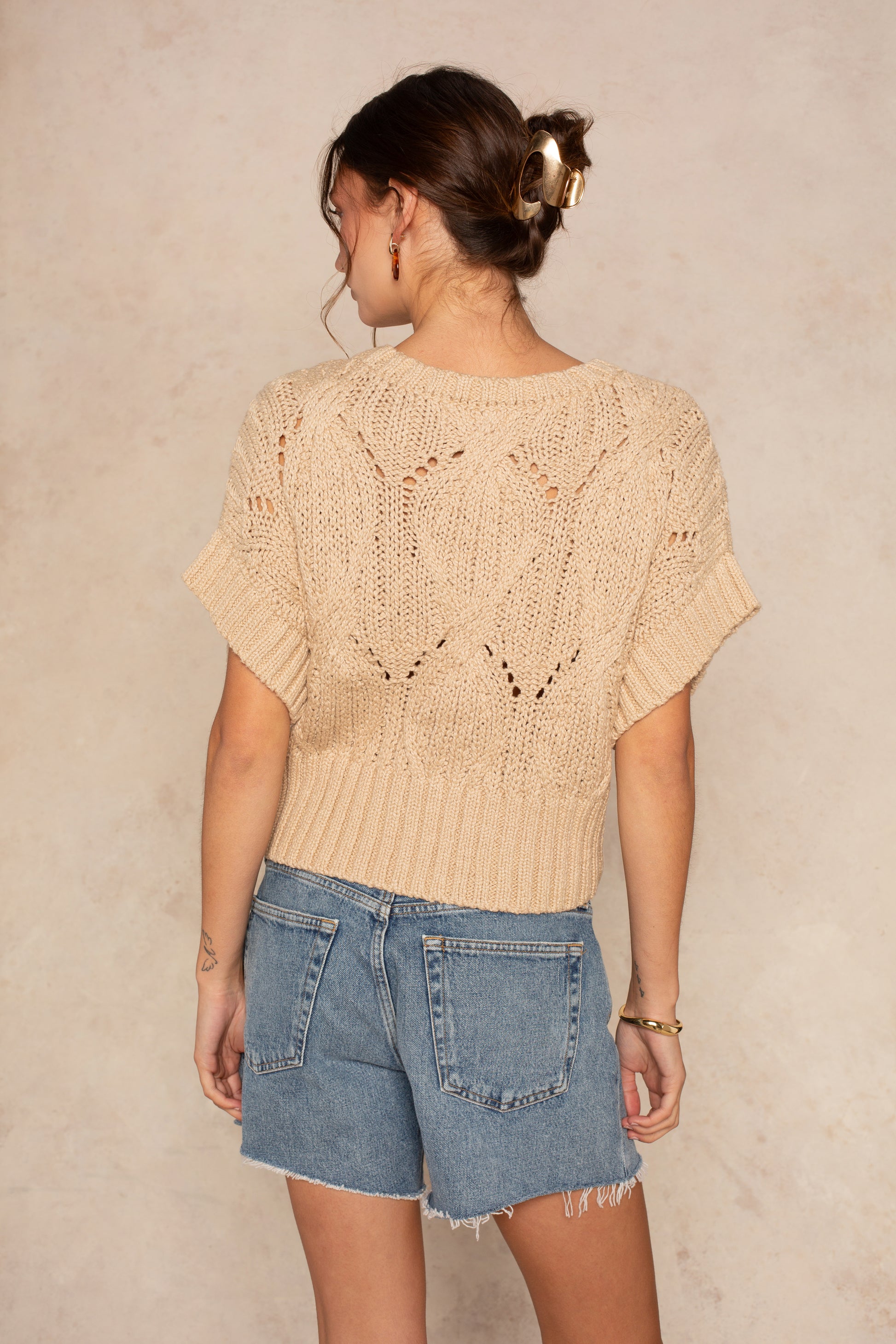 A woman with her hair in a clip wears Isla’s Cali Crochet chunky knit short-sleeve sweater and blue frayed denim shorts, standing with her back to the camera against a neutral background.