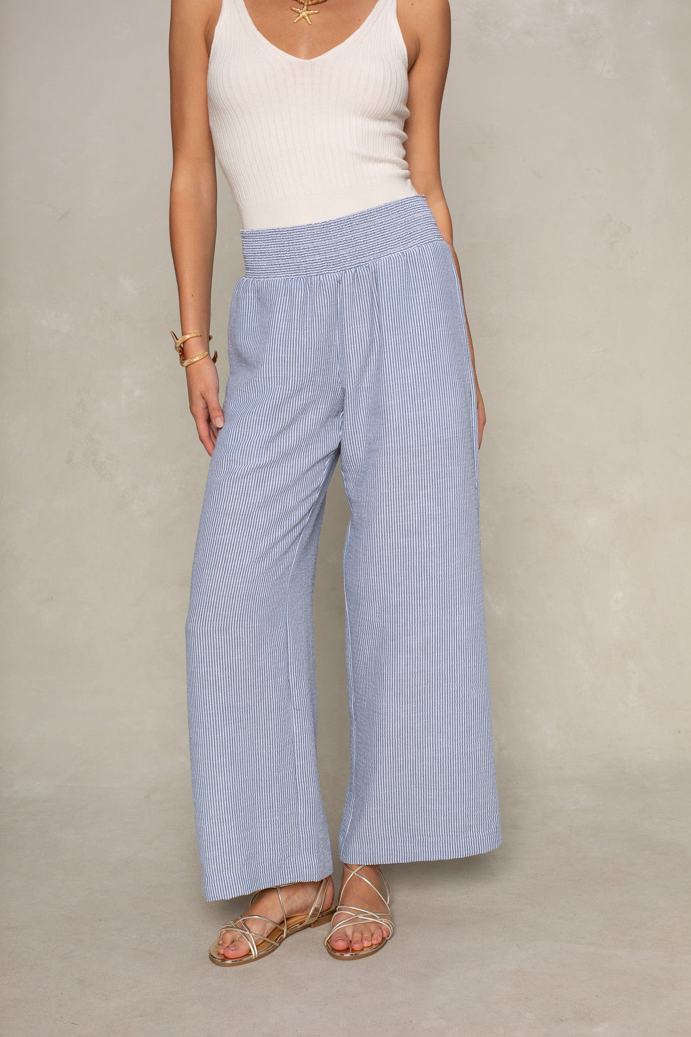 A person models Isla's Hattie Beach Pant—full length, straight leg pants with a smocked waistband—paired with a white sleeveless ribbed top and gold sandals against a neutral background. The person's head is not visible.