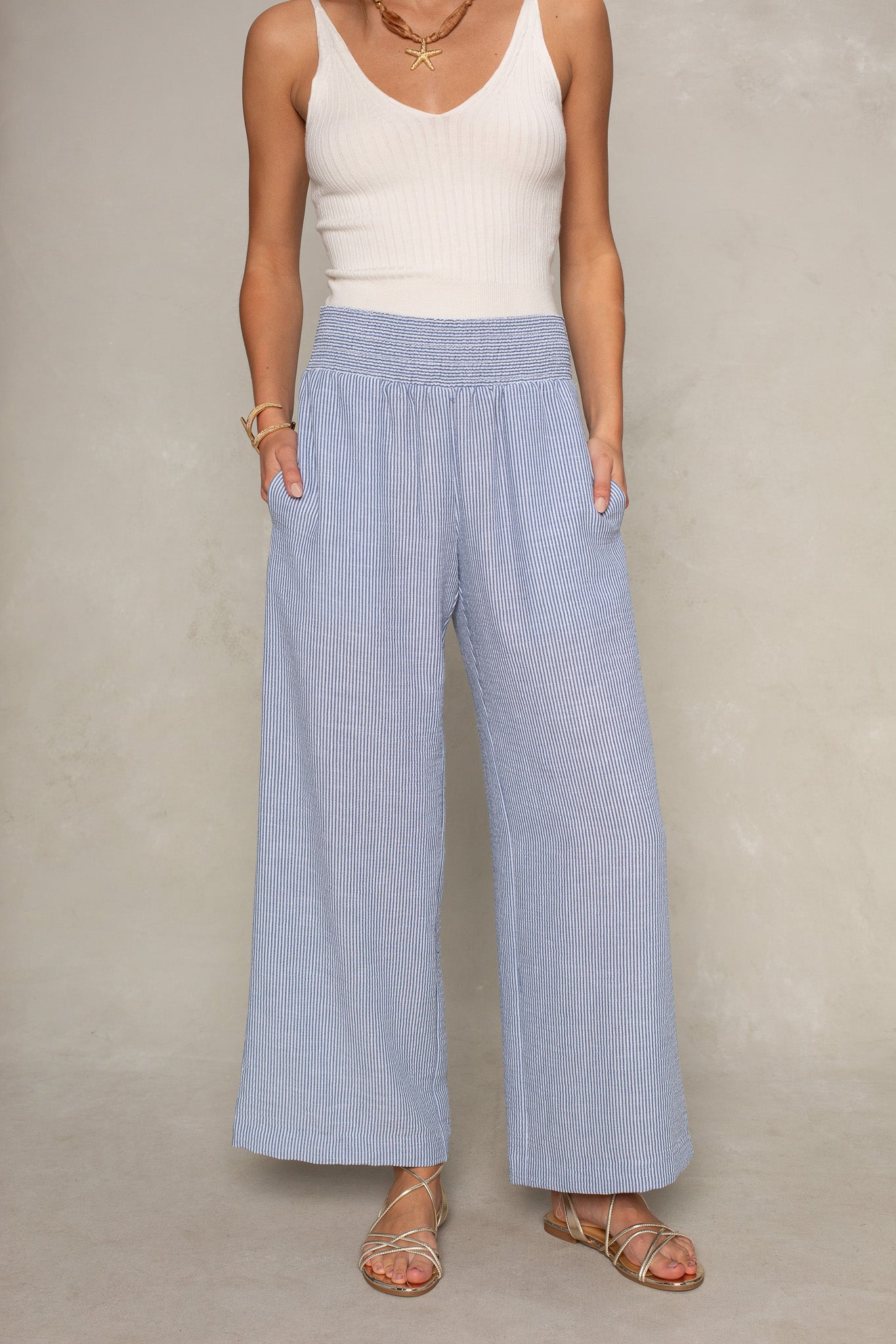 A person wears Isla’s Hattie Beach Pant in navy—a high-waisted, full-length striped style with a smocked waistband and pockets—paired with a sleeveless white ribbed top and gold sandals, standing against a neutral background.