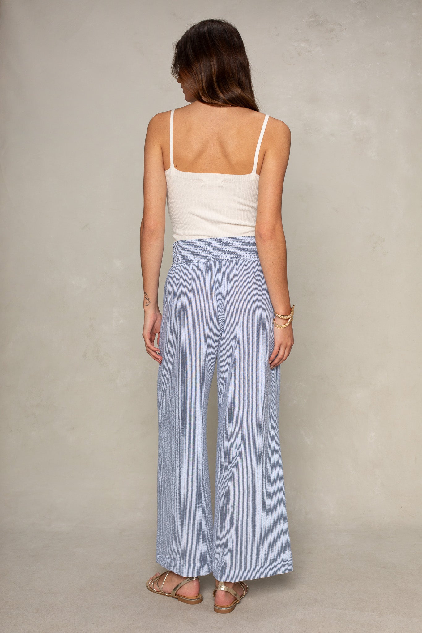 A woman faces away, wearing a white sleeveless top and Isla Hattie Beach Pant—light blue, full-length with a smocked waistband—paired with sandals against a plain, light-colored wall.
