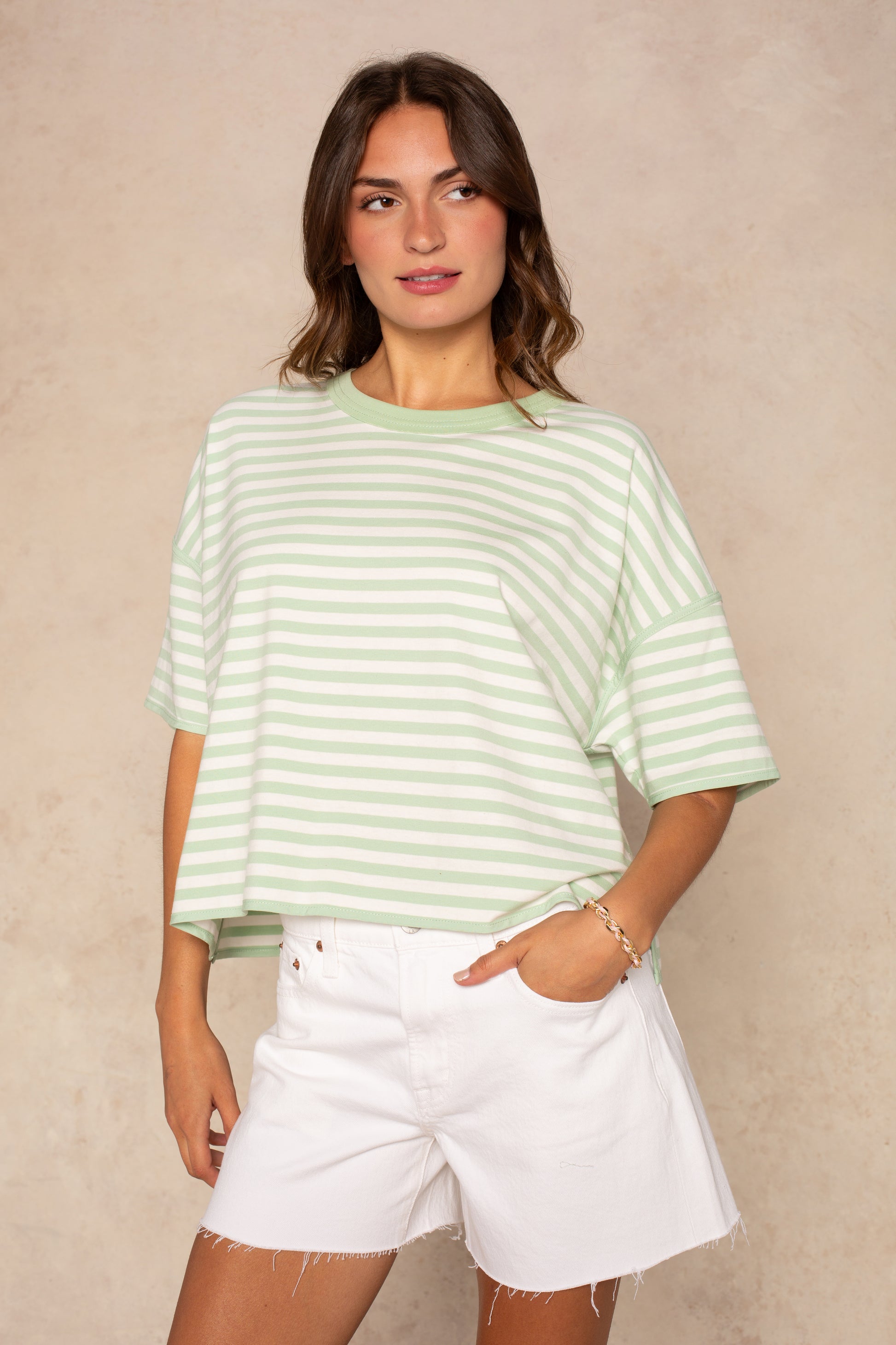 A woman in the Isla Franki Boxy Stripe Knit Tee in Green Multi stands against a beige background, pairing it with white shorts and posing with one hand in her pocket and a relaxed expression.