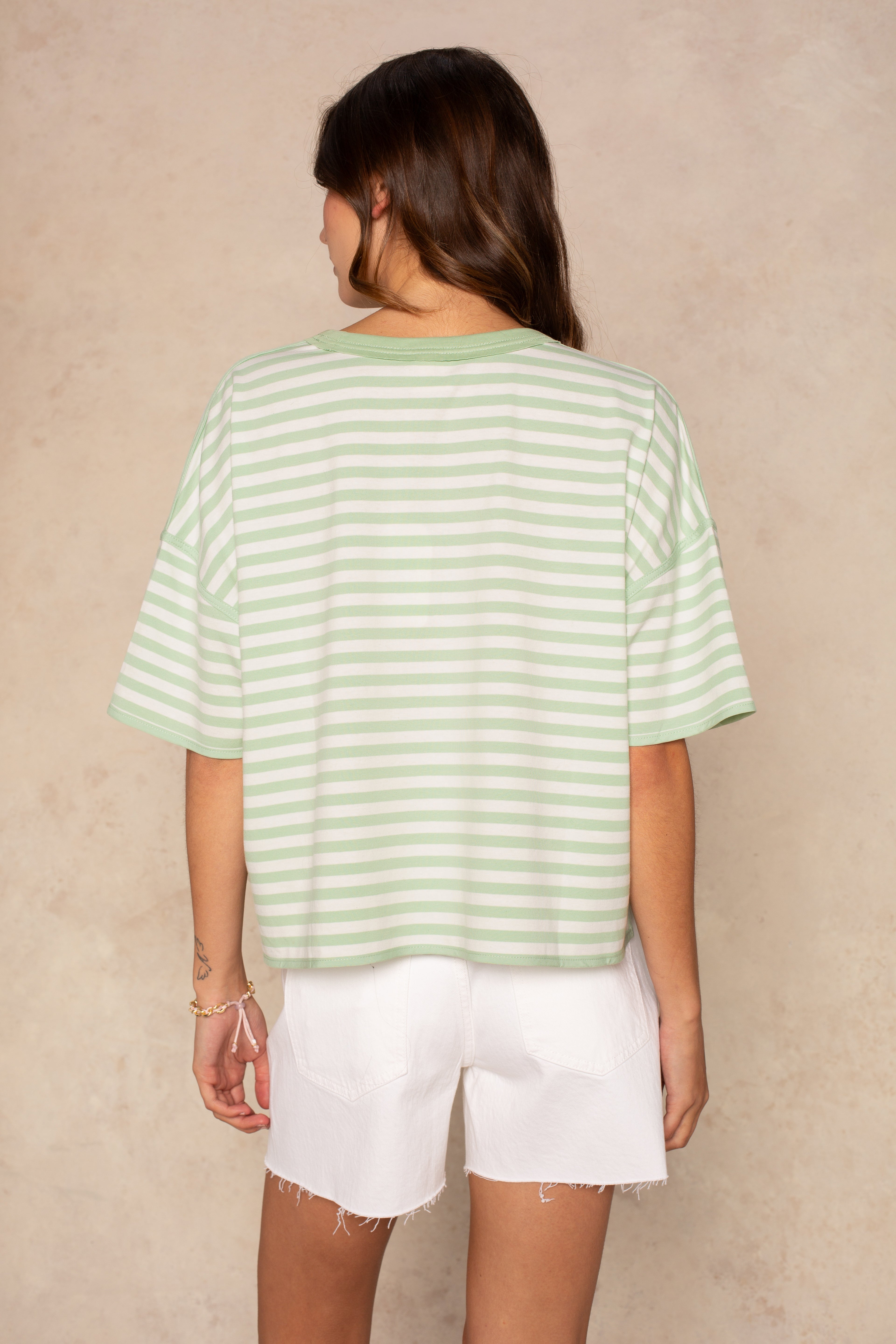 A person with long brown hair, seen from behind, wears the Isla Franki Boxy Stripe Knit Tee in light green and white stripes and white frayed shorts, standing against a beige textured background.