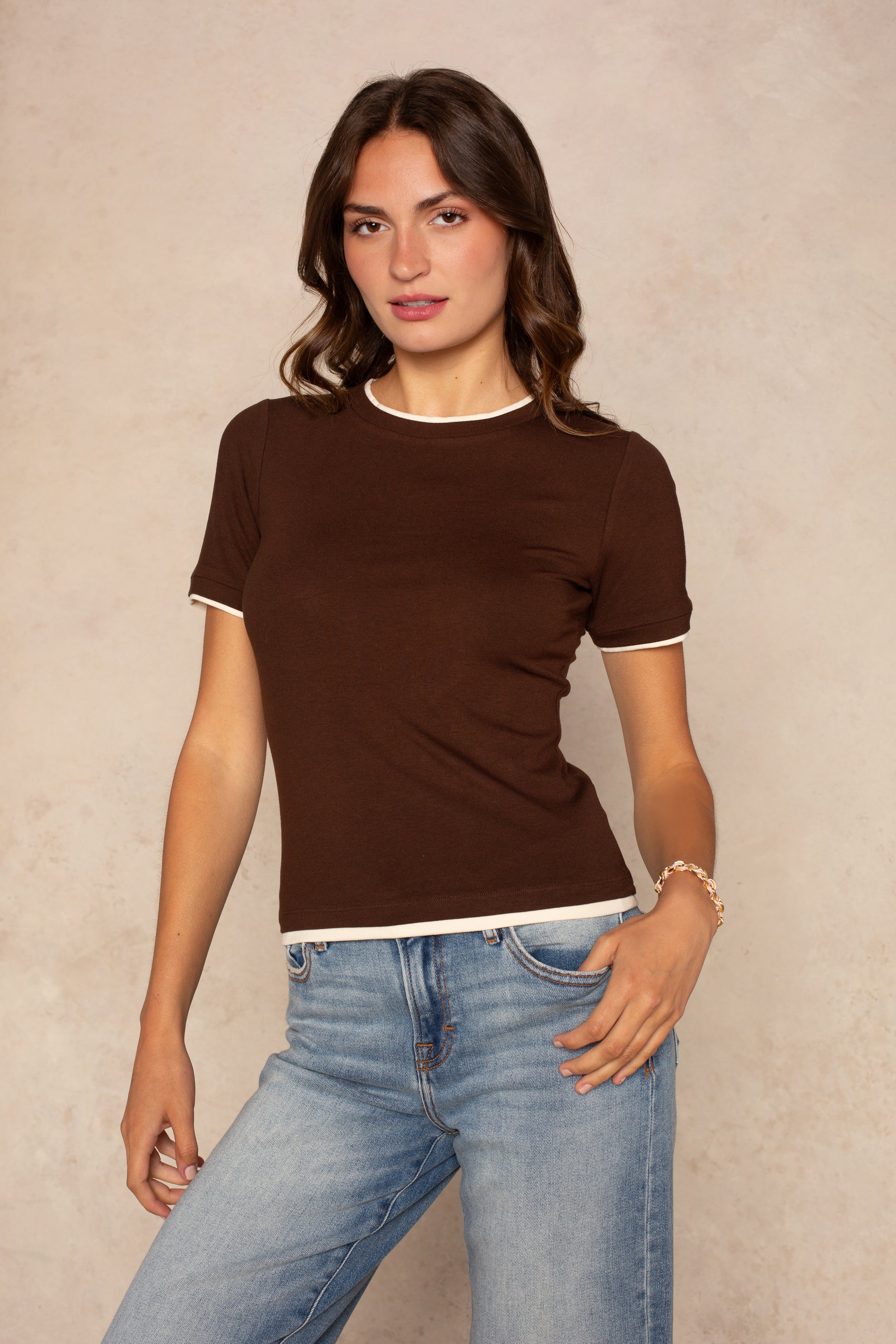A woman with wavy brown hair wears the Isla Jessi Faux Double Tee in Brown Multi and light blue jeans, standing against a neutral beige background.
