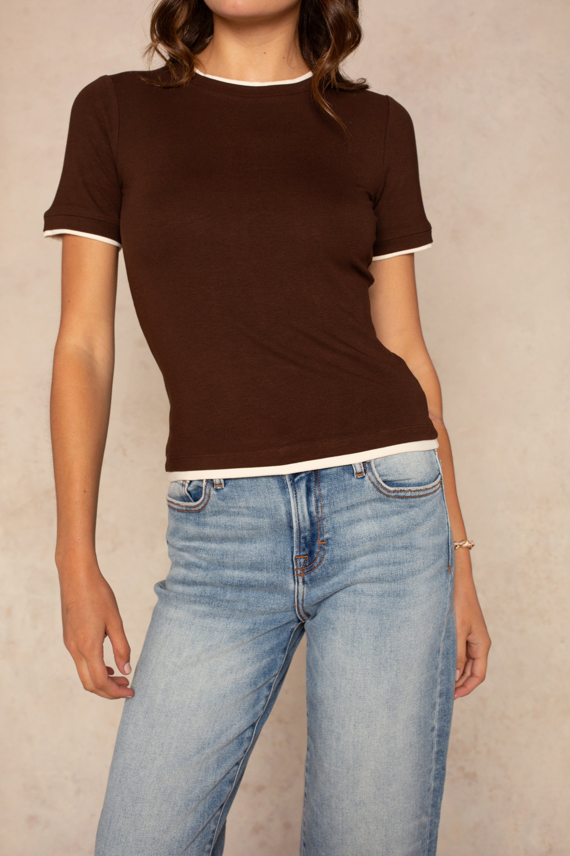 A woman wears the Isla Jessi Faux Double Tee, a vintage-inspired brown fitted baby tee with white contrast trim, paired with light blue jeans. She stands before a beige background, her face partially out of frame.