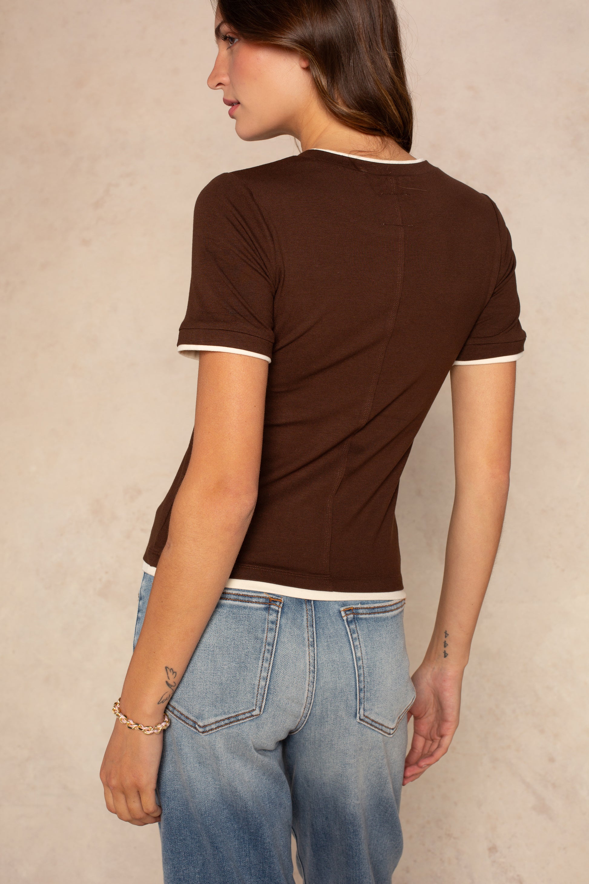 A woman stands with her back to the camera, wearing the Isla Jessi Faux Double Tee—a vintage-inspired brown fitted top with white trim—paired with light blue jeans against a neutral background.