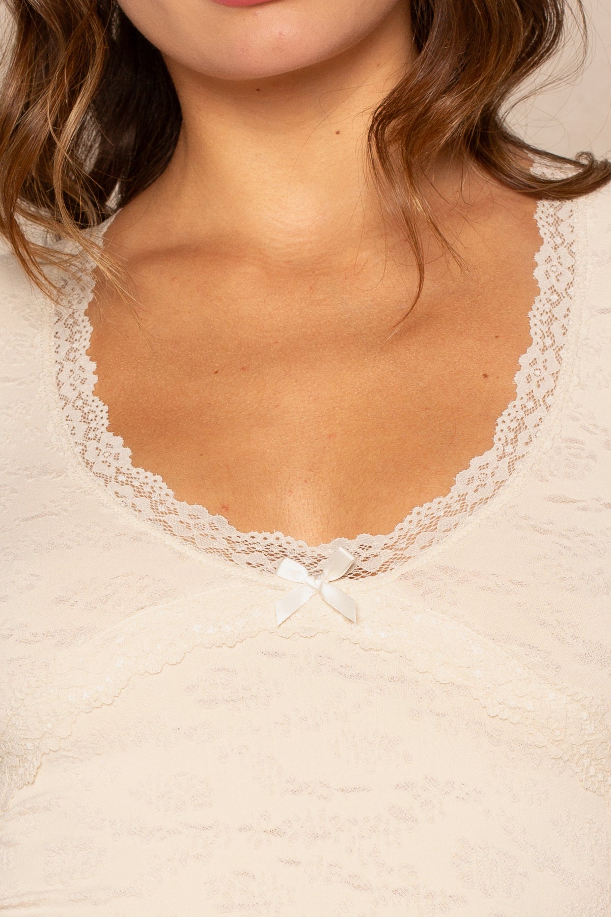 Close-up of a woman wearing the Isla Scoop Neck Lacey Tee, a cream-colored fitted top with lace trim and a small white bow. Her wavy brown hair partially frames her mouth and chin.