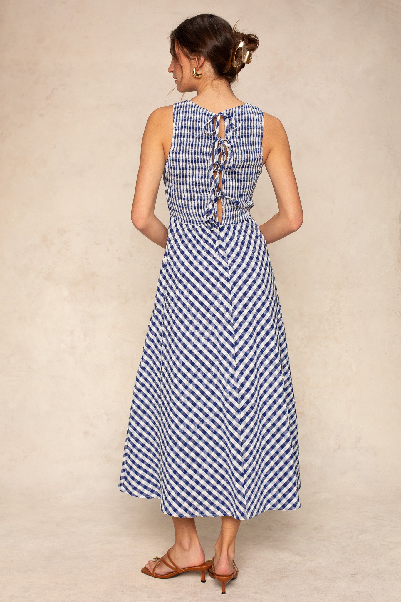 A woman faces away, wearing the Georgie Lola Smocked Gingham Maxi Dress in blue and white with a lace-up back and maxi length. Her hair is up, she wears brown sandals, and a neutral background accentuates the classic gingham style.