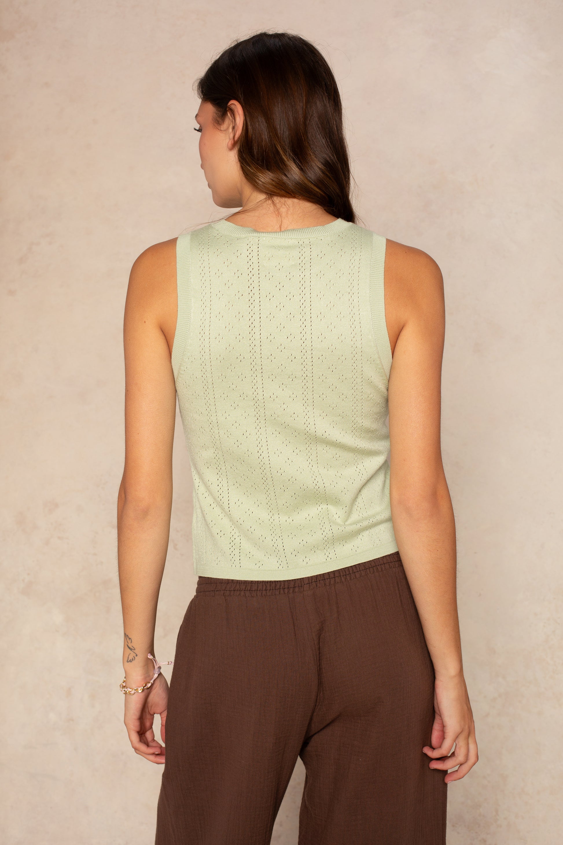 A woman with long brown hair stands facing away, wearing the Isla Anna Sleeveless Pointelle Top in sage green and loose brown pants. She has a small tattoo on her left arm and a bracelet. The background is light beige.