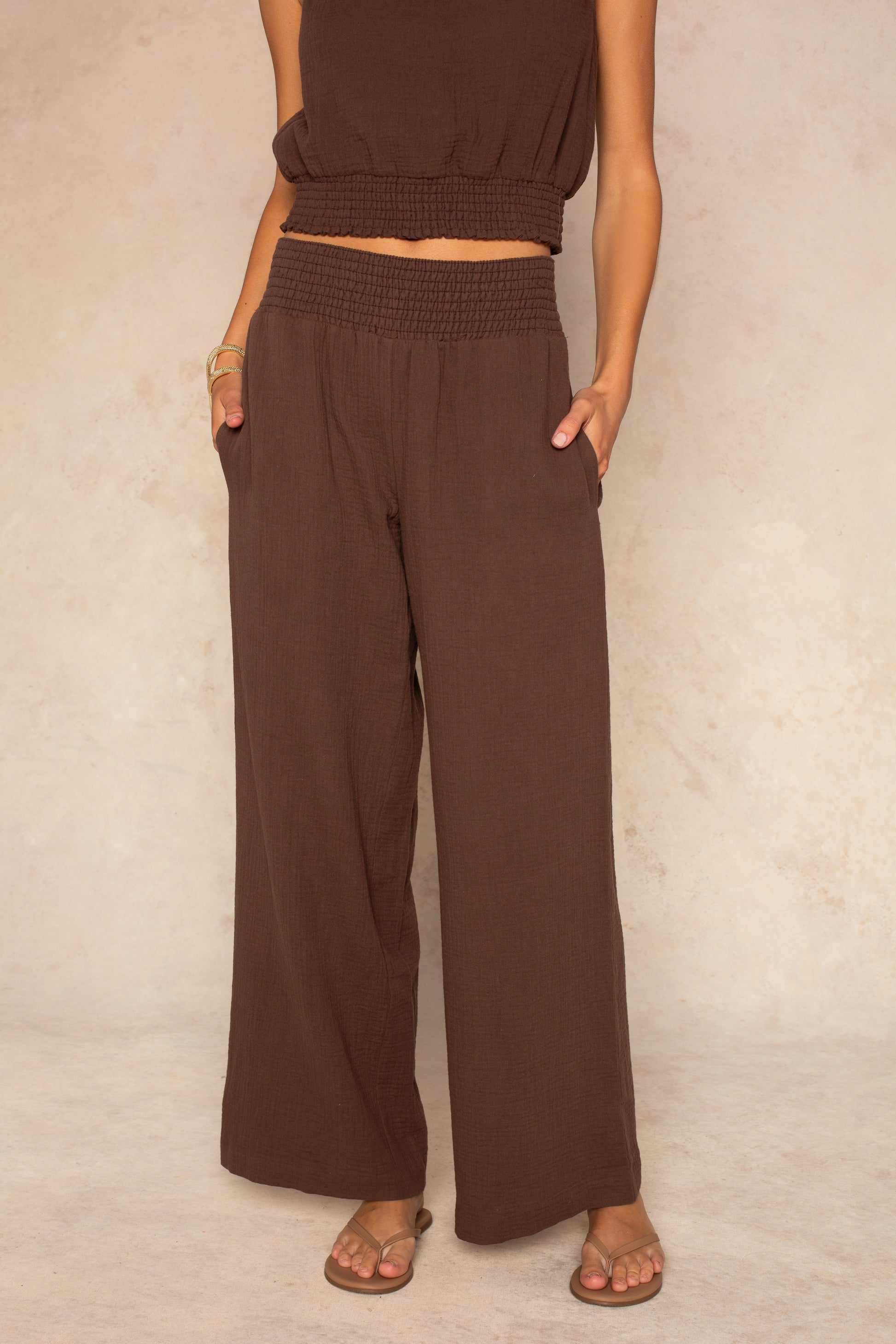 A person wears the Isla Hattie Beach Pant in brown, full length with a smocked waistband and straight legs, paired with a matching brown sleeveless crop top and sandals. Their hands are in their pockets; face not visible, standing on a neutral floor.