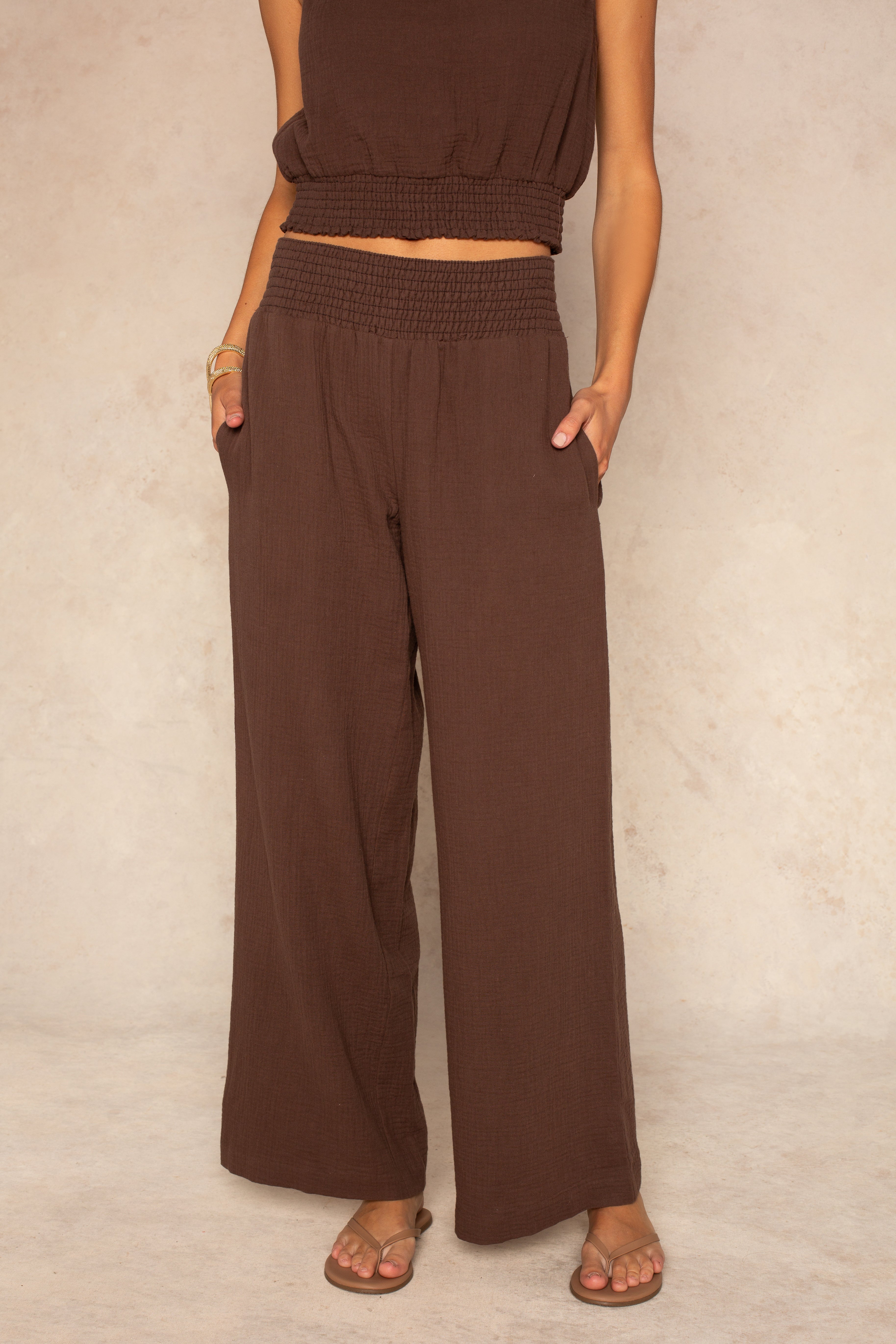 A person wears the Isla Hattie Beach Pant in brown, full length with a smocked waistband and straight legs, paired with a matching brown sleeveless crop top and sandals. Their hands are in their pockets; face not visible, standing on a neutral floor.