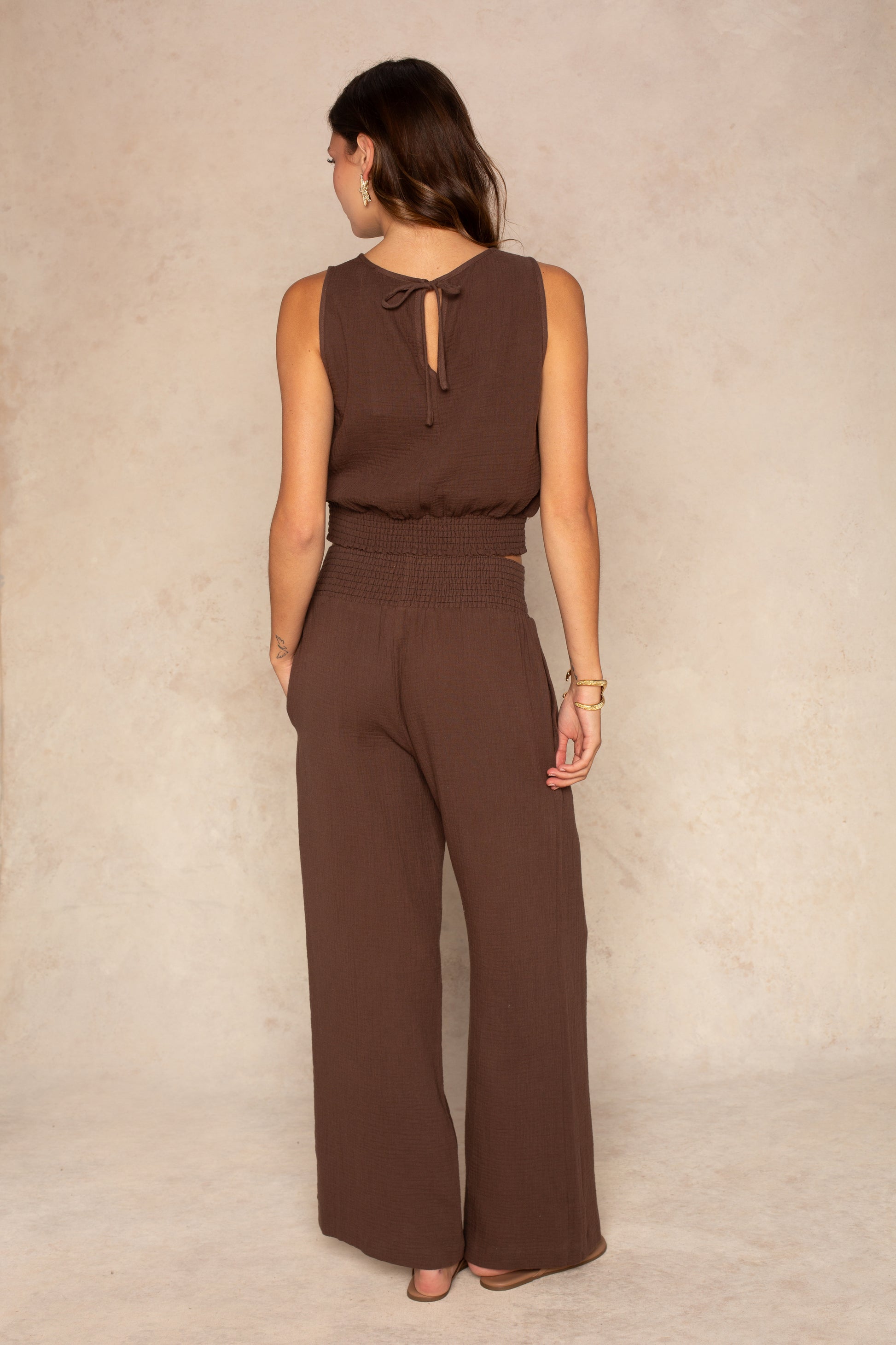 A woman with brown hair stands with her back to the camera, wearing the Isla Harley Gauze Sleeveless Top. She accessorizes with hoop earrings and bracelets.