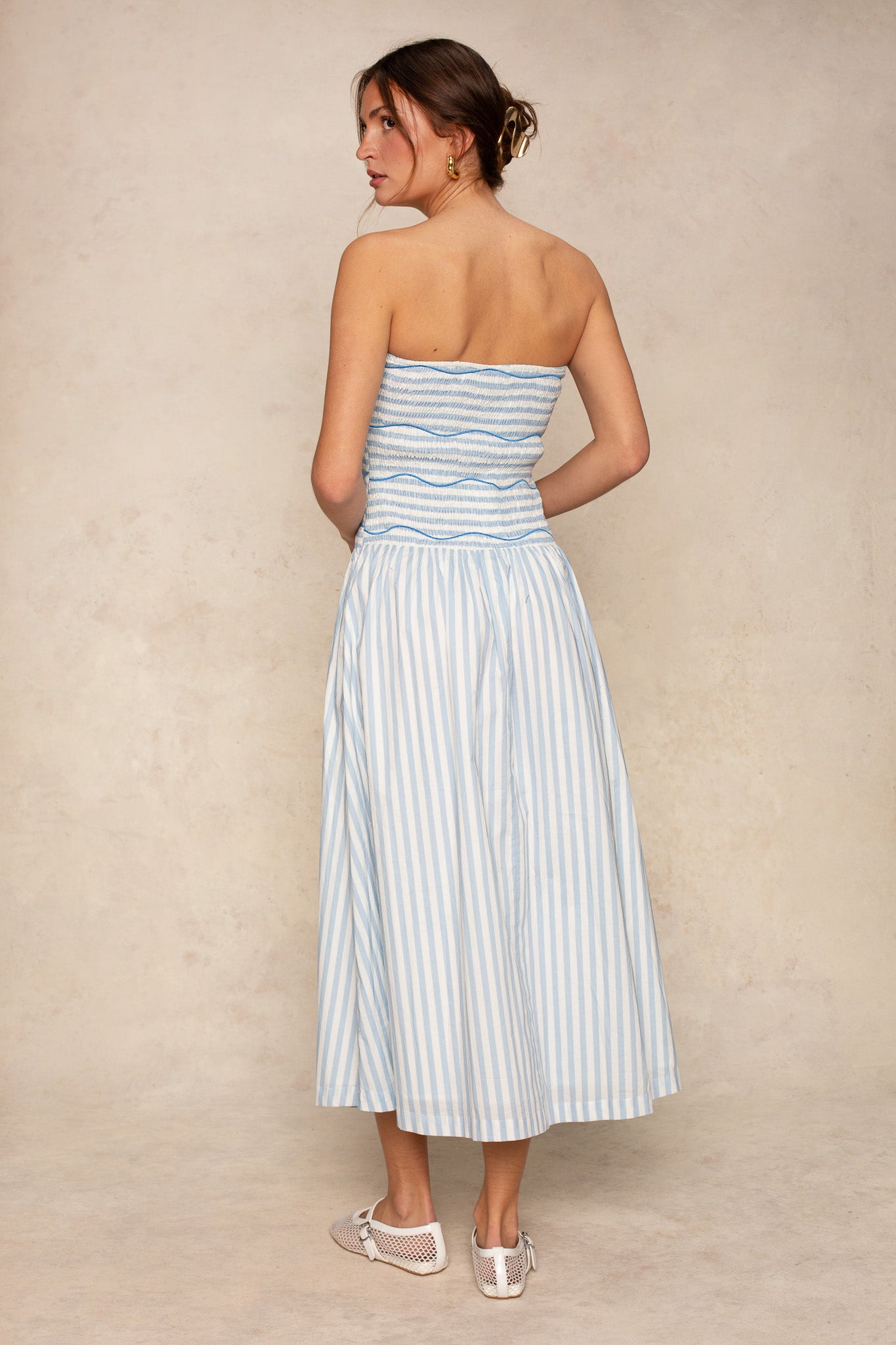 A woman faces away, wearing the Georgie Nora Strapless Smocked Midi Dress in light blue and white stripes. Her hair is up, styled with gold hoop earrings and white mesh flats for a versatile, chic look.