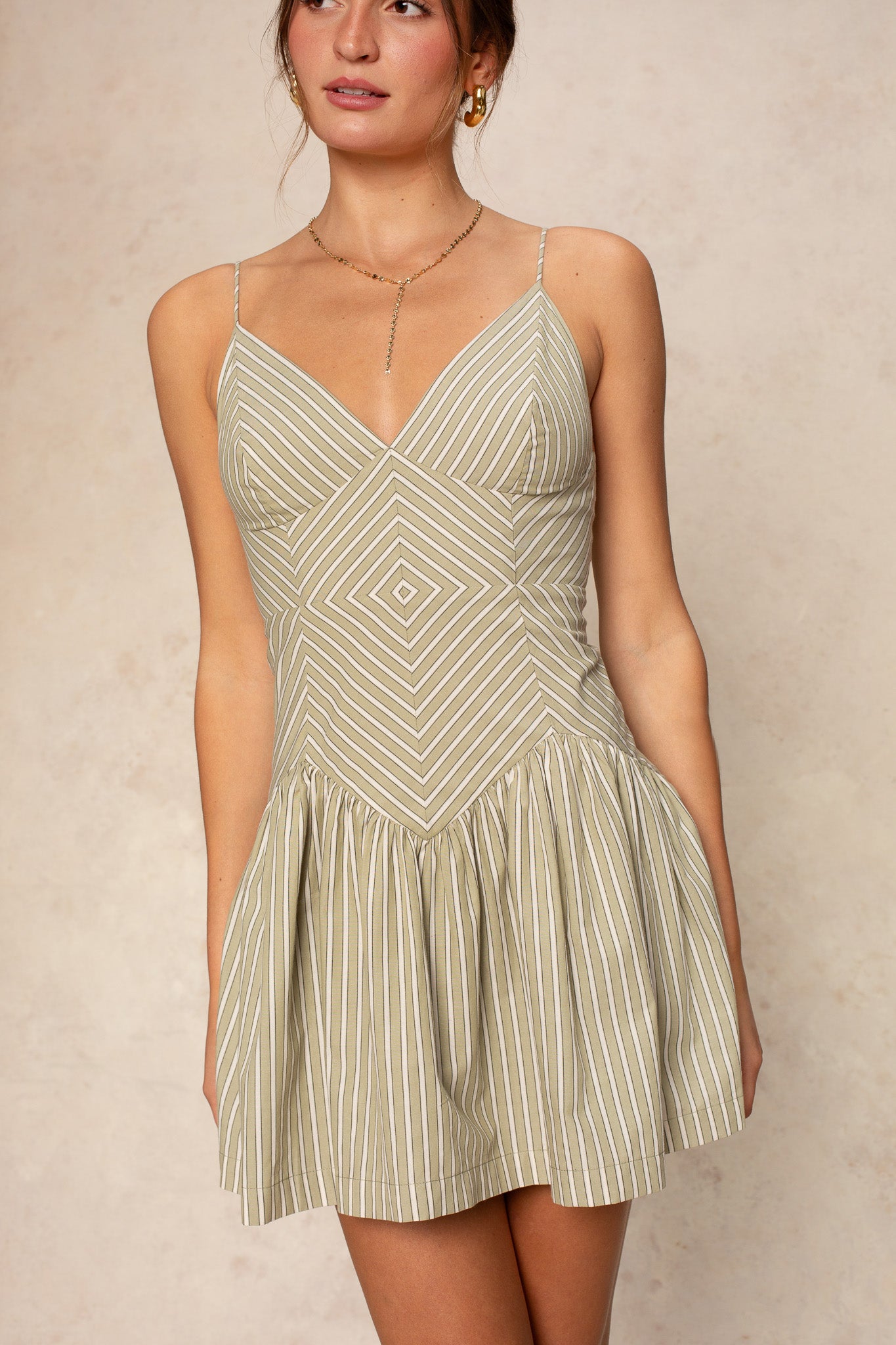 A woman wears the Giselle Stripe Mini by Georgie, a light beige and white striped spaghetti strap dress with a fitted bodice, flared skirt, and geometric patterns. Her hair is tied back and she accessorizes with a necklace.