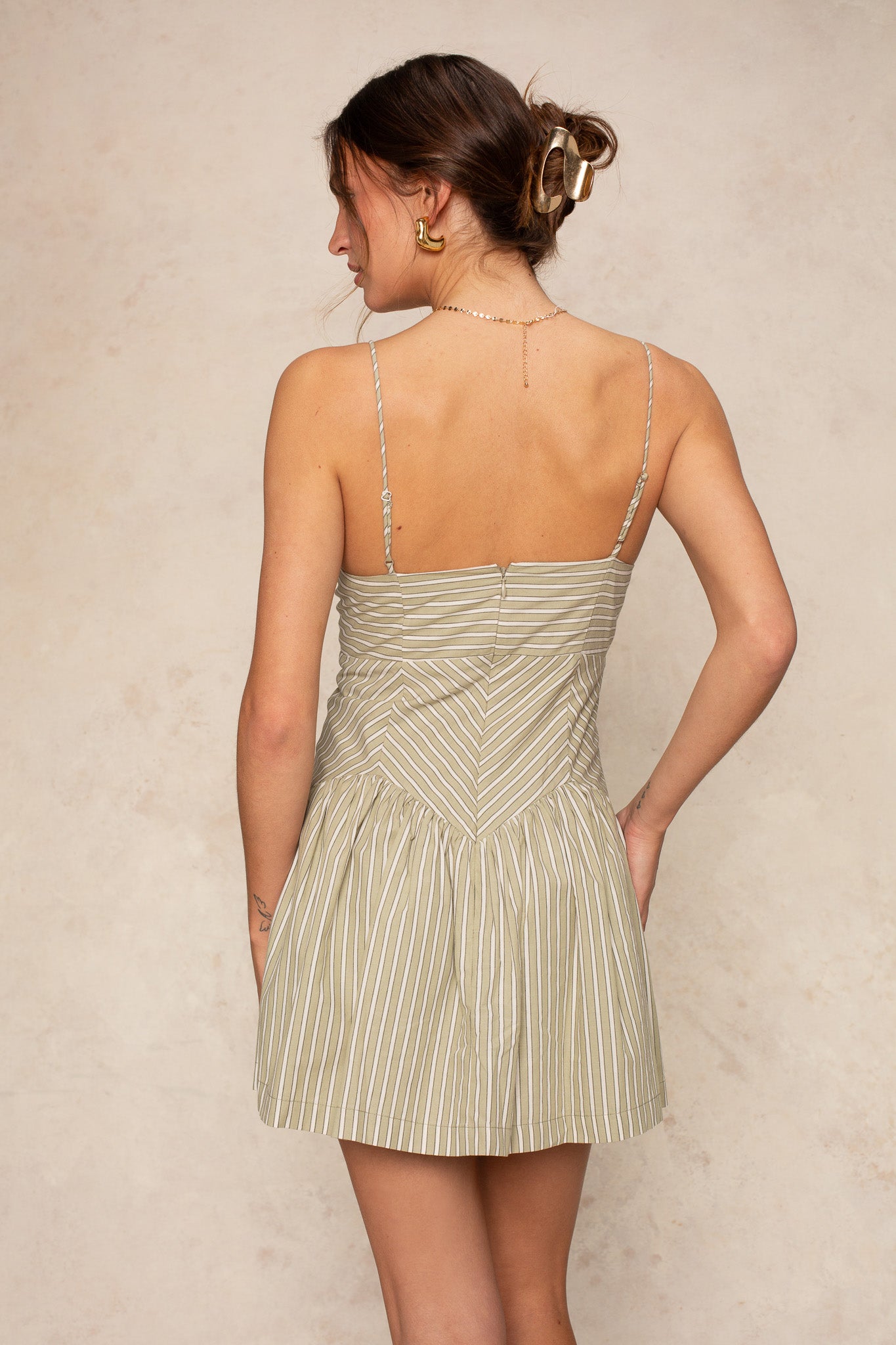 A woman with brown hair in a loose bun, wearing gold earrings and the Georgie Giselle Stripe Mini sundress with thin green and white straps, stands with her back to the camera against a beige background, radiating timeless sophistication.