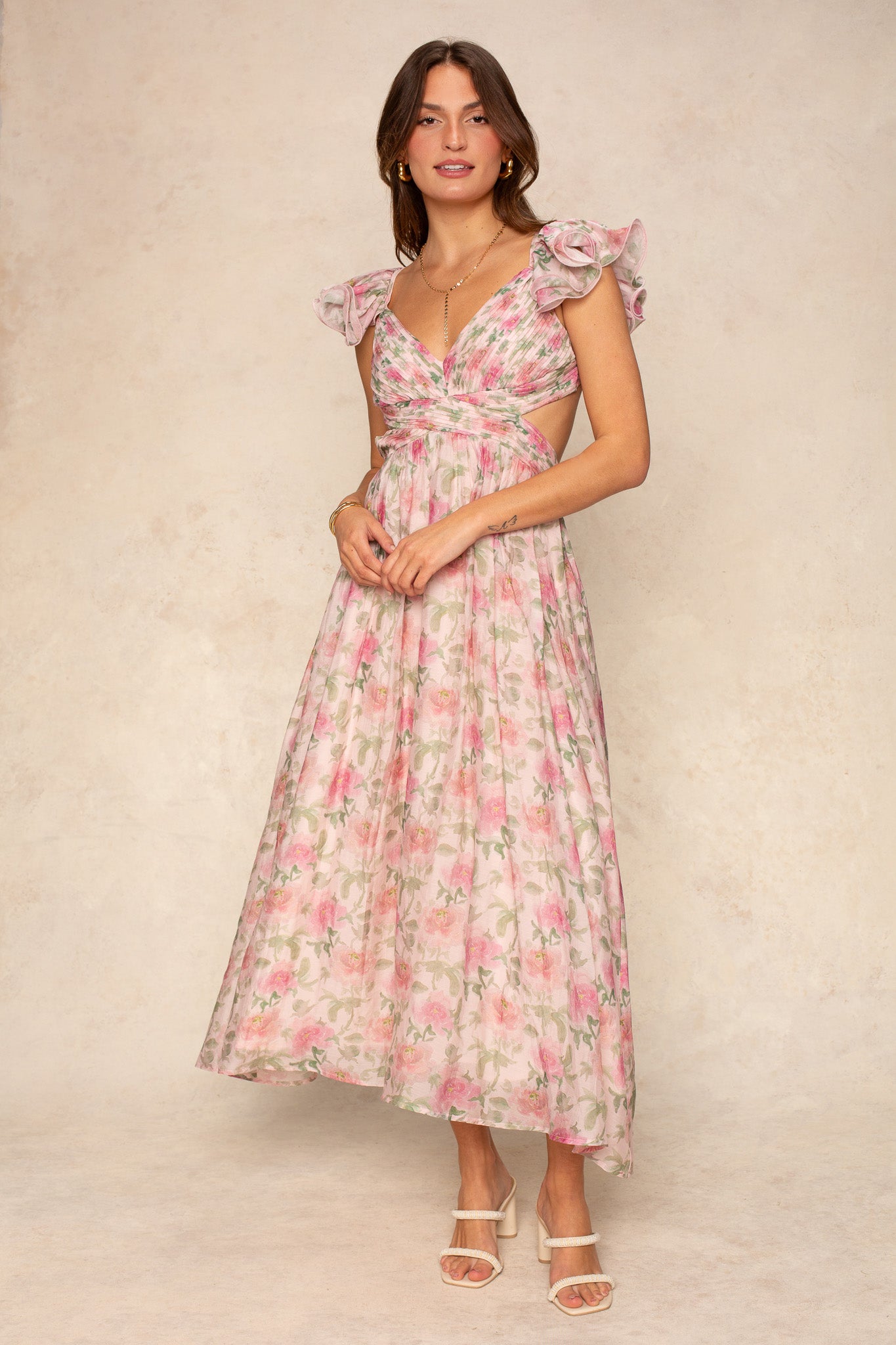 A woman wears the Georgie Rosalie Flutter Sleeve Maxi Dress in light pink floral print, featuring flutter sleeves, a gathered bodice, and an under-bust cutout. She pairs it with layered necklaces and white heels against a neutral backdrop.