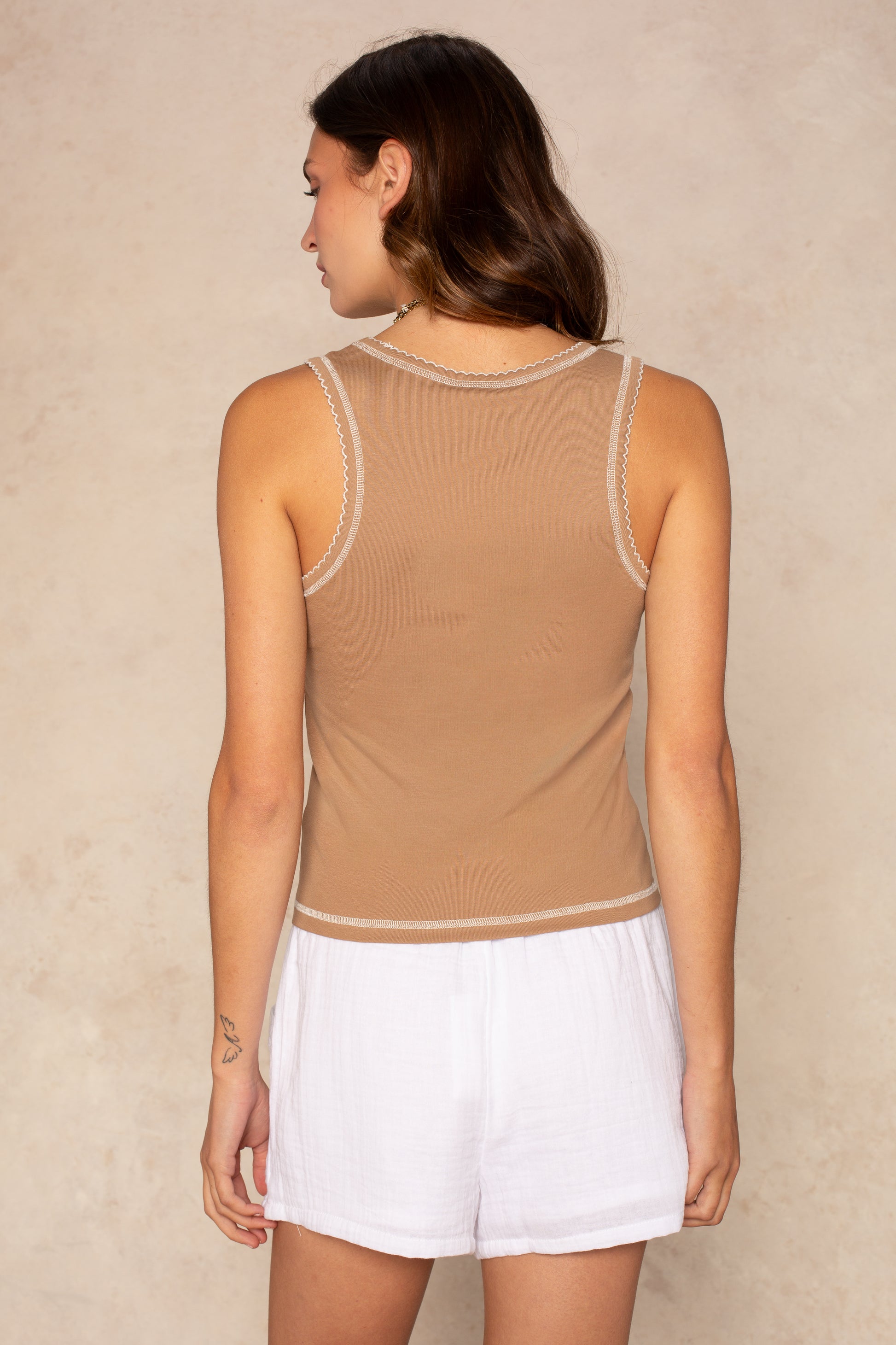 A woman with long brown hair, seen from behind, wears the Isla Tess Double Trim Stitch Tank in tan with white trim and loose white shorts, standing against a neutral beige wall.
