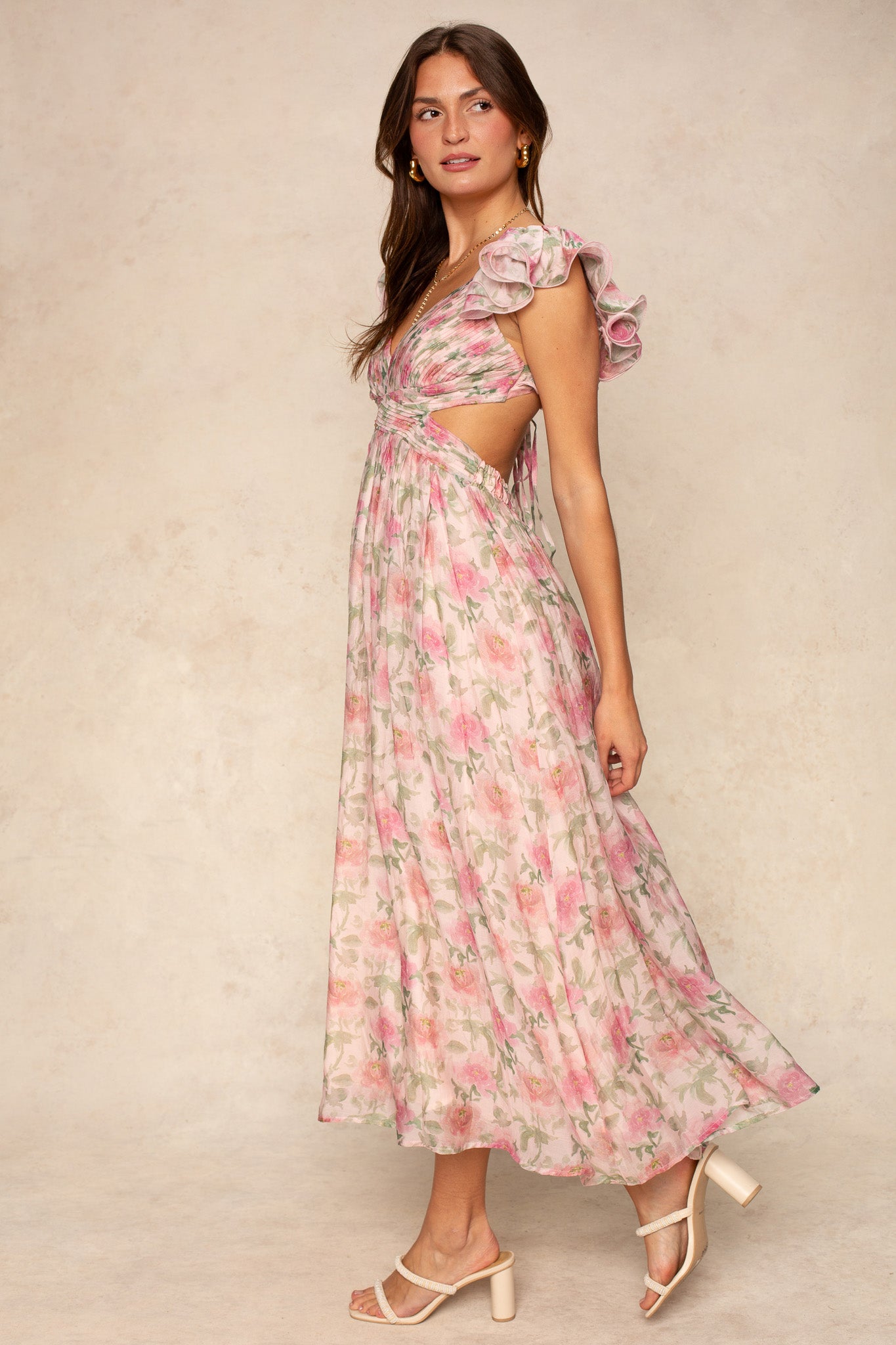 A woman stands sideways in the Georgie Rosalie Flutter Sleeve Maxi Dress—a flowy, floral pink and green special occasion gown with flutter sleeves and side cutouts—paired with open-toe cream heels against a neutral background.