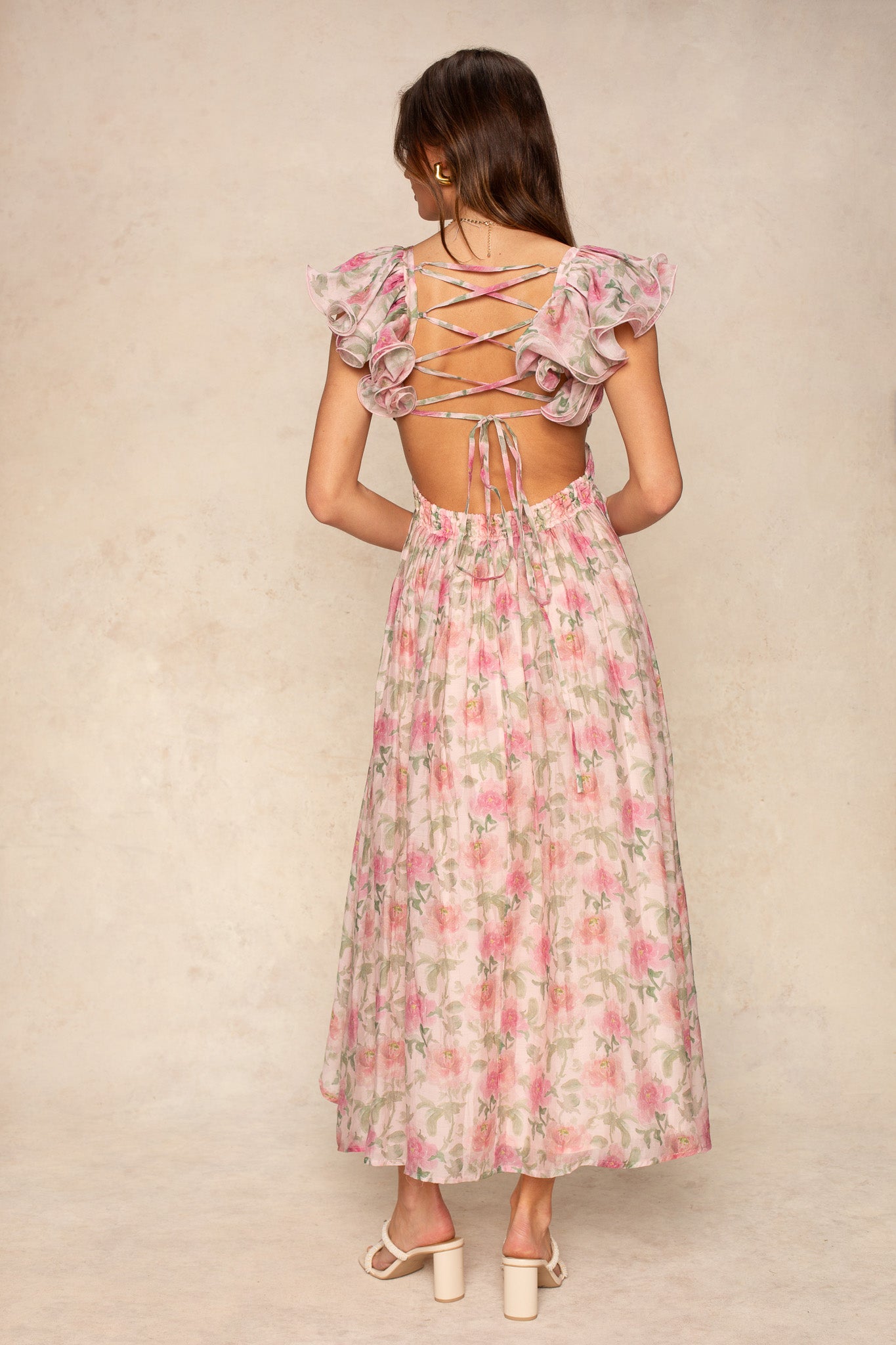 A woman faces away in the Georgie Rosalie Flutter Sleeve Maxi Dress—a pink floral maxi with flutter sleeves and a lace-up open back. Her long brown hair flows down, paired with white heeled sandals—ideal for special occasions.