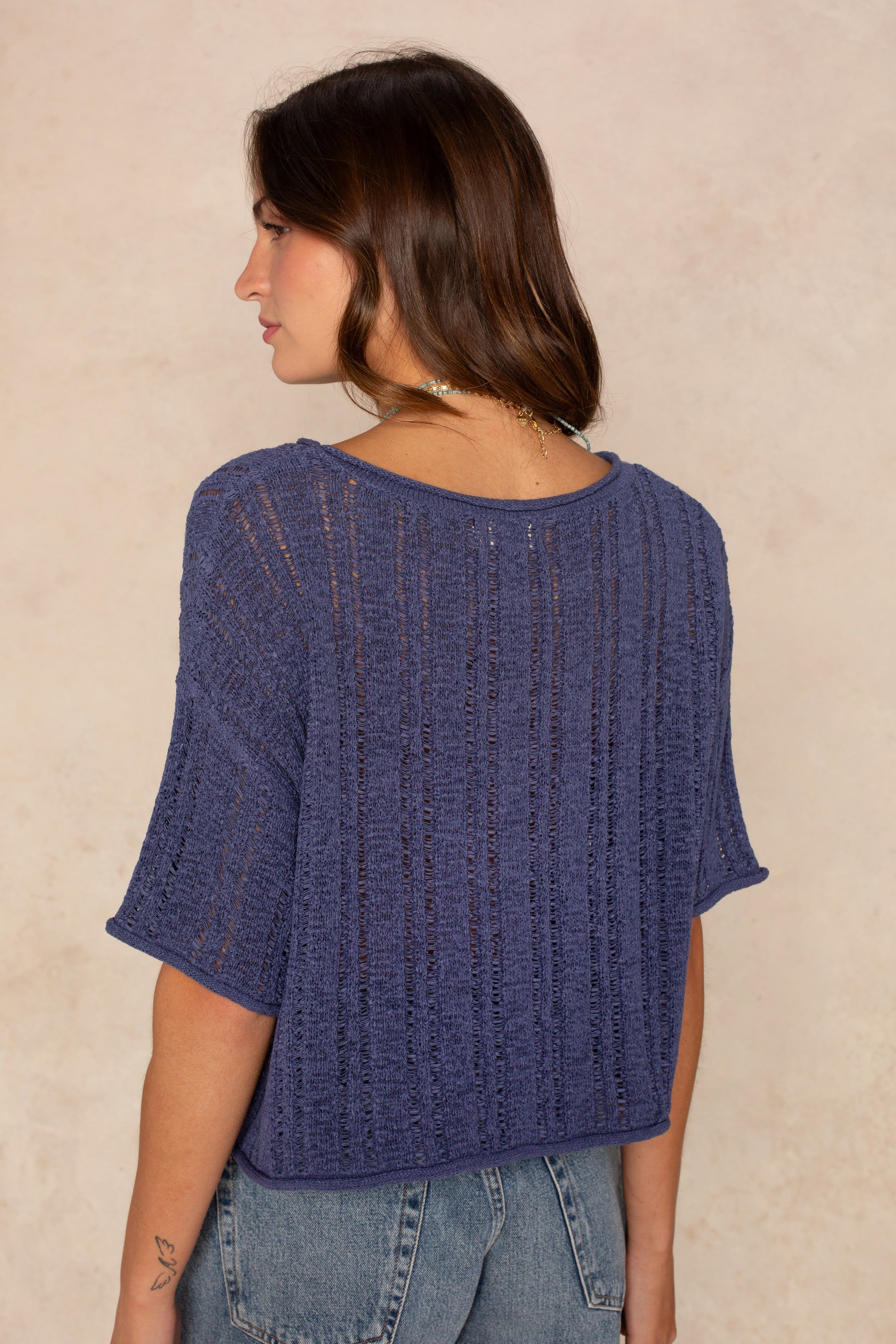 A woman with shoulder-length brown hair stands with her back to the camera, wearing Isla’s Brielle Crochet Short Sleeve Crew Neck in blue and light blue jeans. The neutral beige background highlights this perfect summer top.