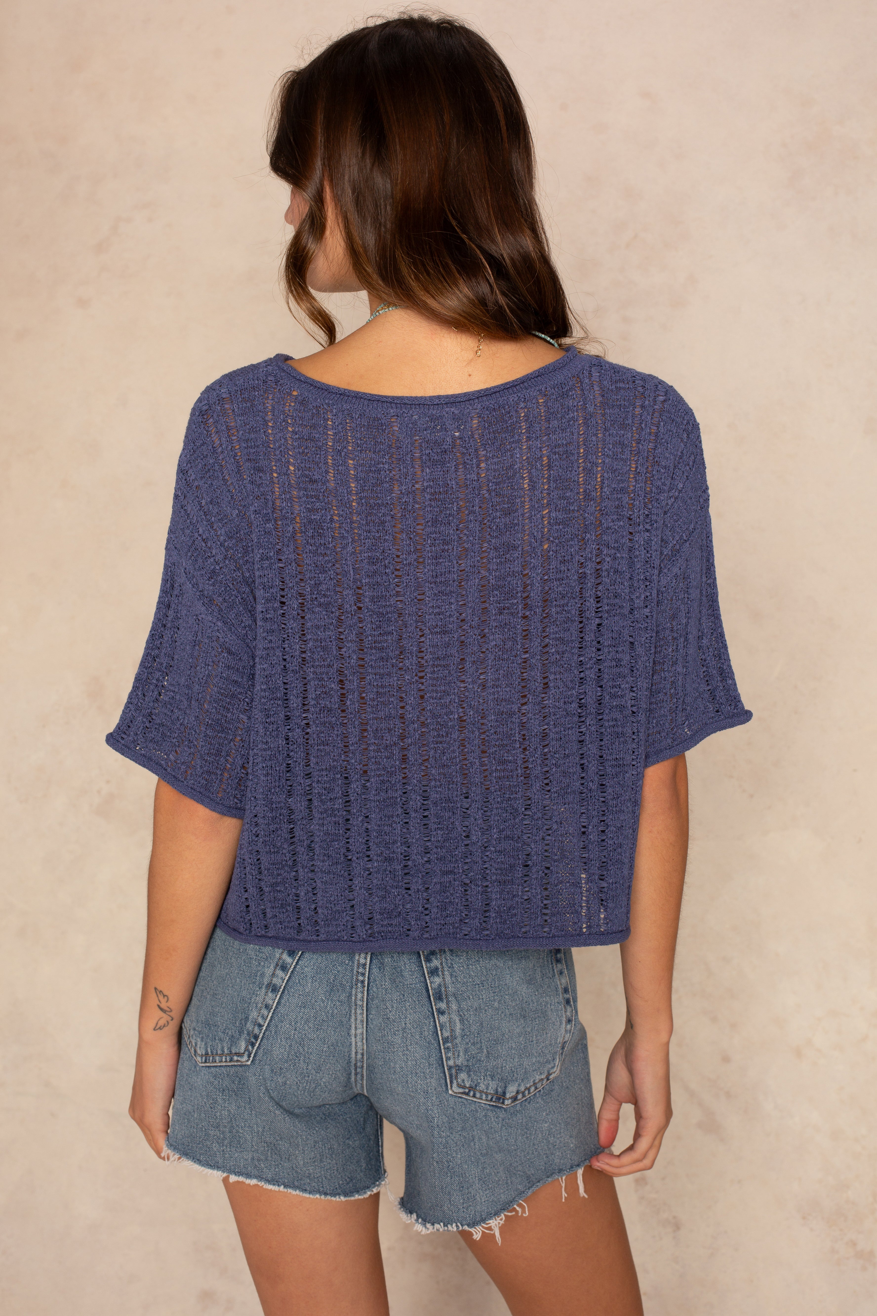 A woman with wavy brown hair stands with her back to the camera, wearing the Isla Brielle Crochet Short Sleeve Crew Neck in purple and high-waisted light blue denim shorts against a neutral beige background.