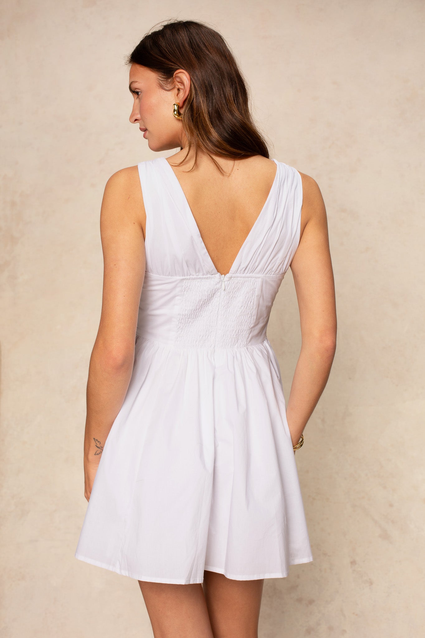 A woman with long brown hair wears the Georgie Vivianne Corset Mini Dress, a sleeveless white piece featuring a deep V-back and ruched, fitted waist. She stands facing away, showcasing the striking back detail against a neutral background.