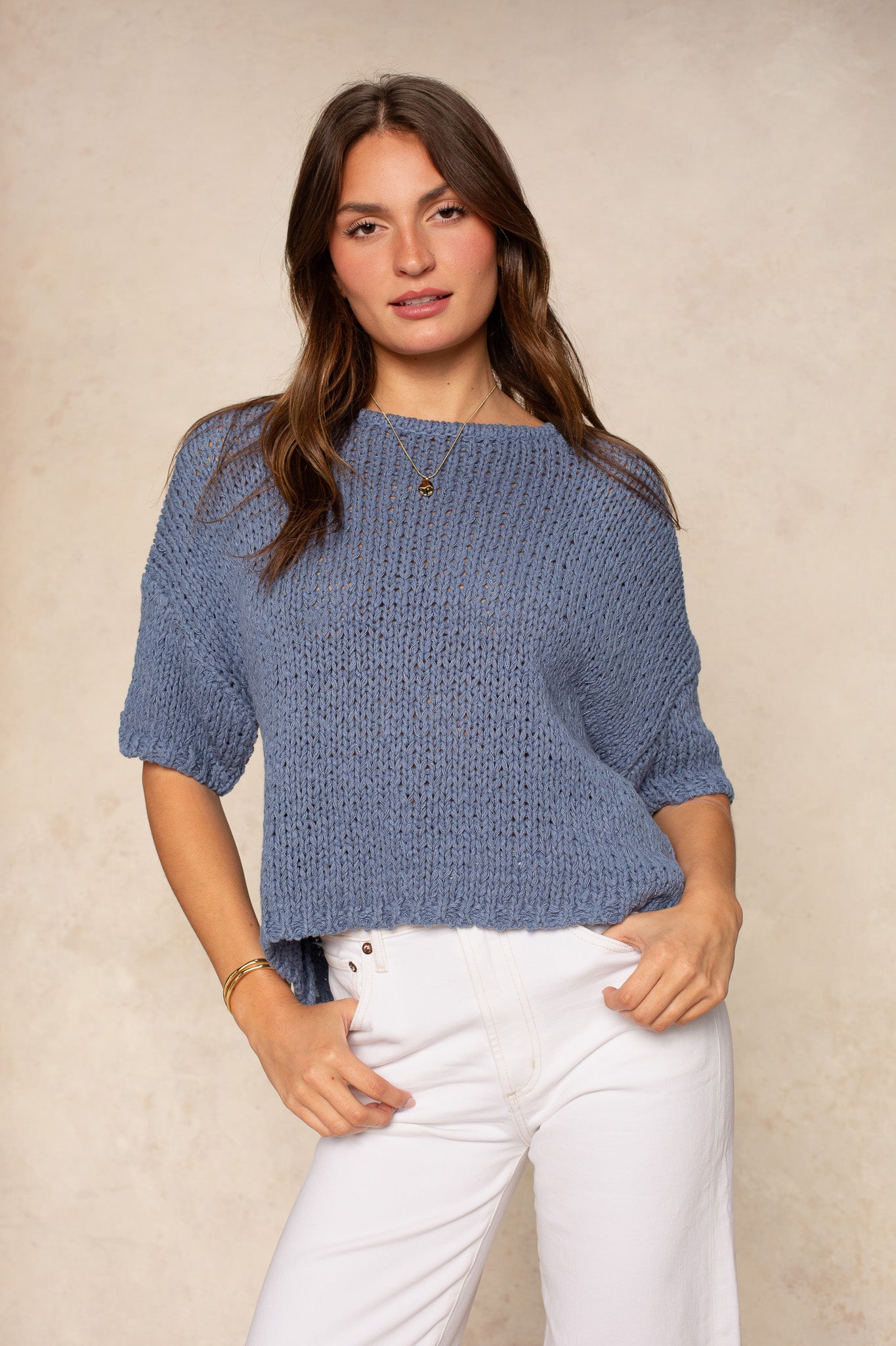 A woman wears the Georgie Penny Short Sleeve Crew Neck Sweater in navy with high-waisted white pants, standing against a beige background—an effortless wardrobe staple.