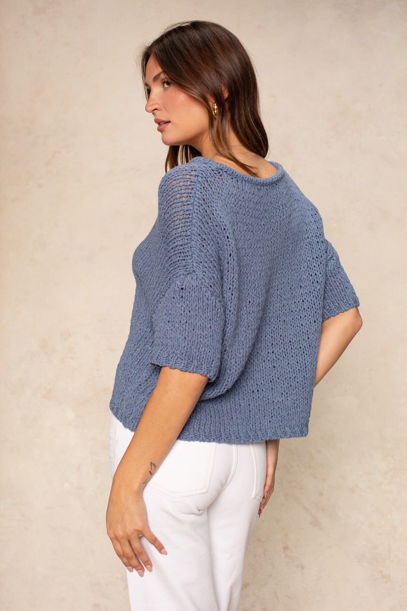 A woman with long brown hair stands sideways against a beige background, wearing Georgie's Penny Short Sleeve Crew Neck Sweater in blue and white pants. She looks over her shoulder with a relaxed expression.