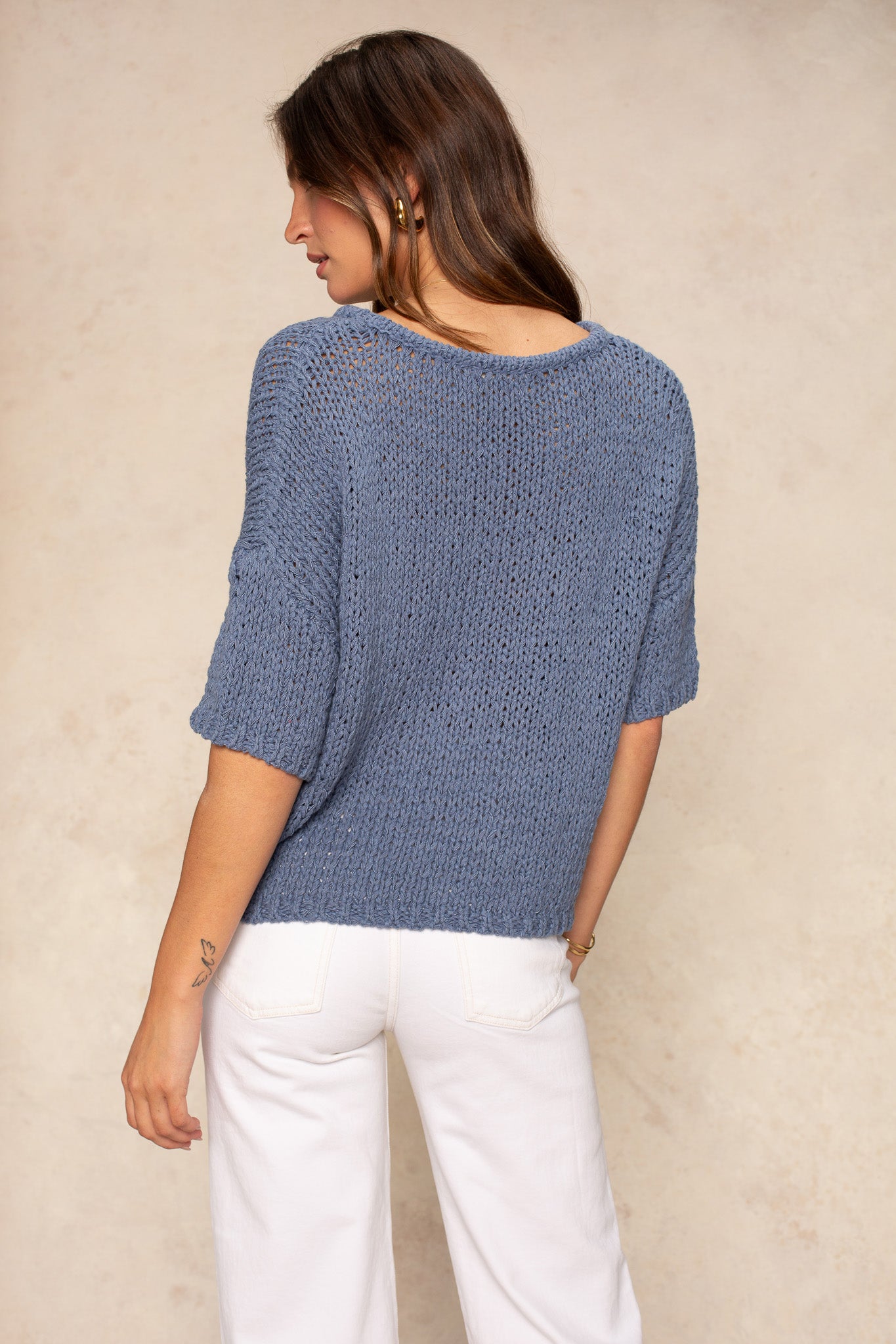 A woman with long brown hair stands with her back to the camera, wearing Georgie’s Penny Short Sleeve Crew Neck Sweater in blue and white pants against a beige background—a true wardrobe staple.