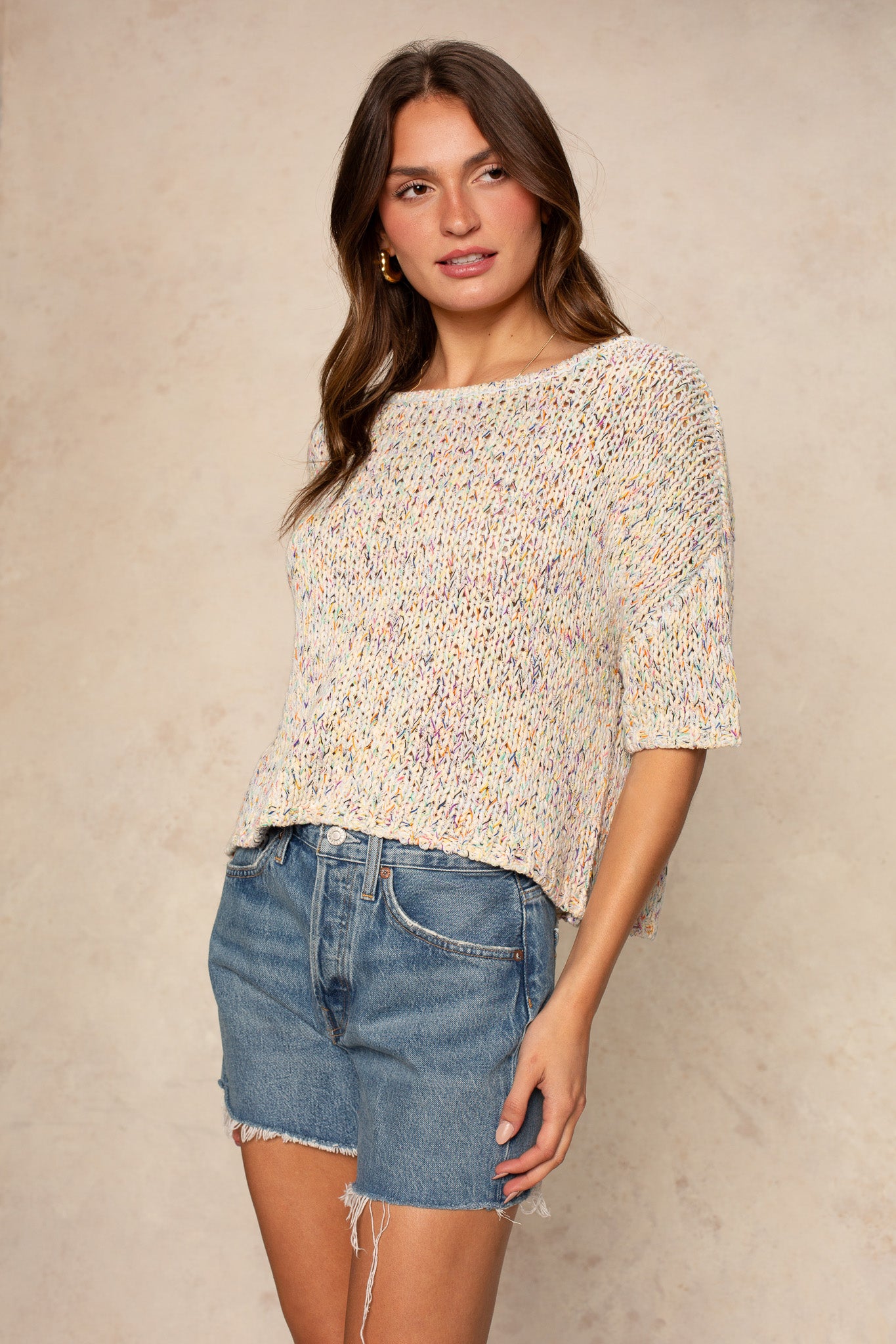 A woman wears the Georgie Penny Short Sleeve Crew Neck Sweater with high-waisted, frayed denim shorts, standing against a neutral background and gazing to the side with a relaxed look.