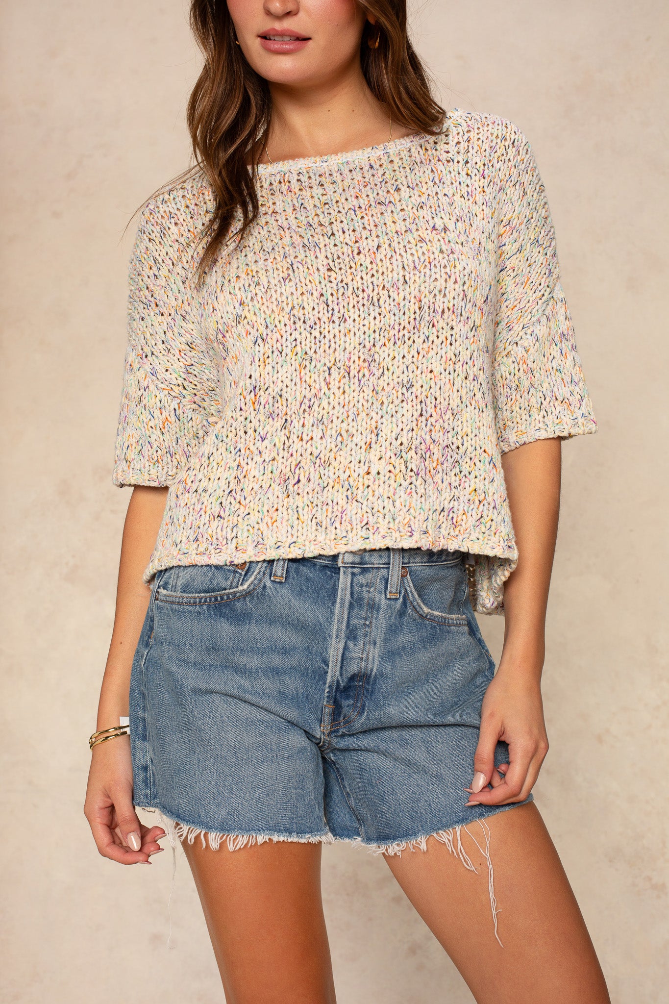 A woman wears the Georgie Penny Short Sleeve Crew Neck Sweater, a light multicolored knit with elbow-length sleeves, paired with high-waisted frayed denim shorts, standing against a neutral beige background.