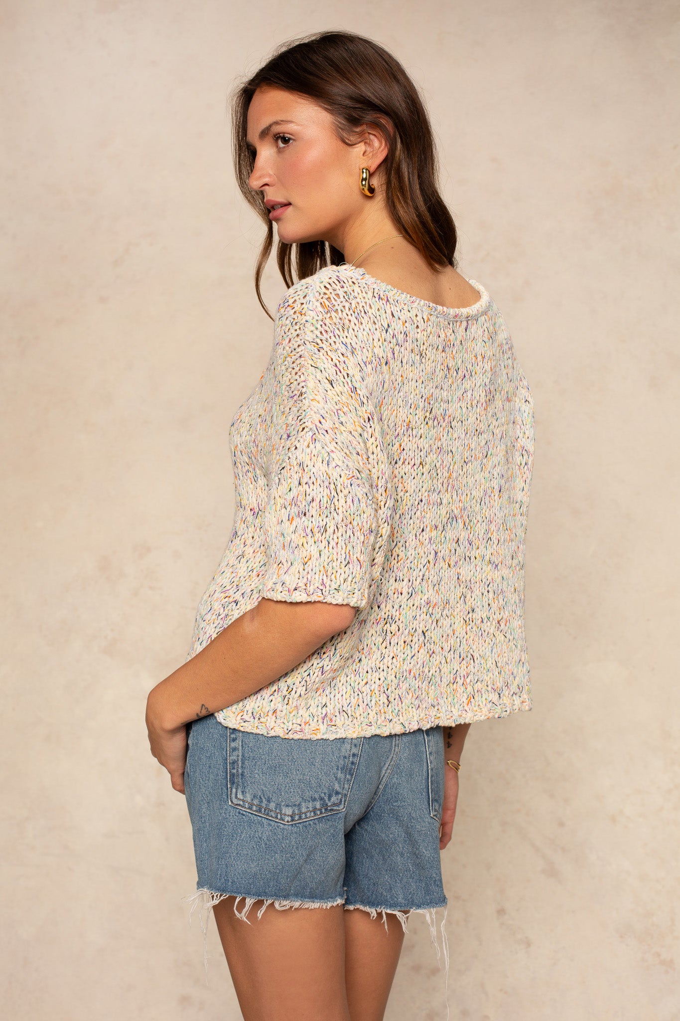 A woman with long brown hair and gold hoop earrings poses sideways against a neutral background, wearing the Georgie Penny Short Sleeve Crew Neck Sweater in a relaxed multicolored knit, paired with light blue denim shorts—a true wardrobe staple.