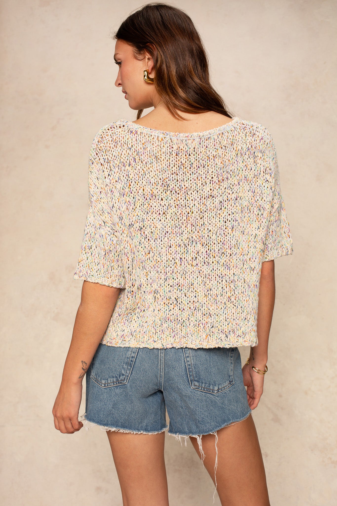 A woman with long brown hair wears the Georgie Penny Short Sleeve Crew Neck Sweater and frayed denim shorts, standing with her back to a beige background.