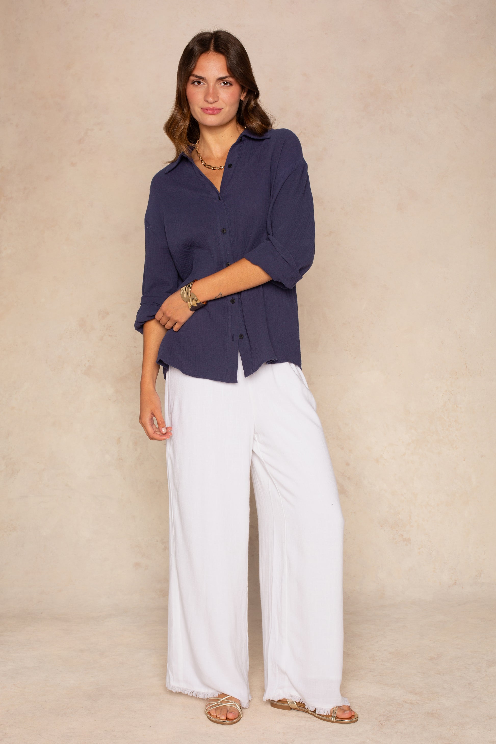 A woman with wavy brown hair, smiling slightly, stands against a beige background wearing the Isla Gia Gauze Button Down Shirt Coverup in navy, styled with white wide-leg pants, sandals, and gold jewelry.
