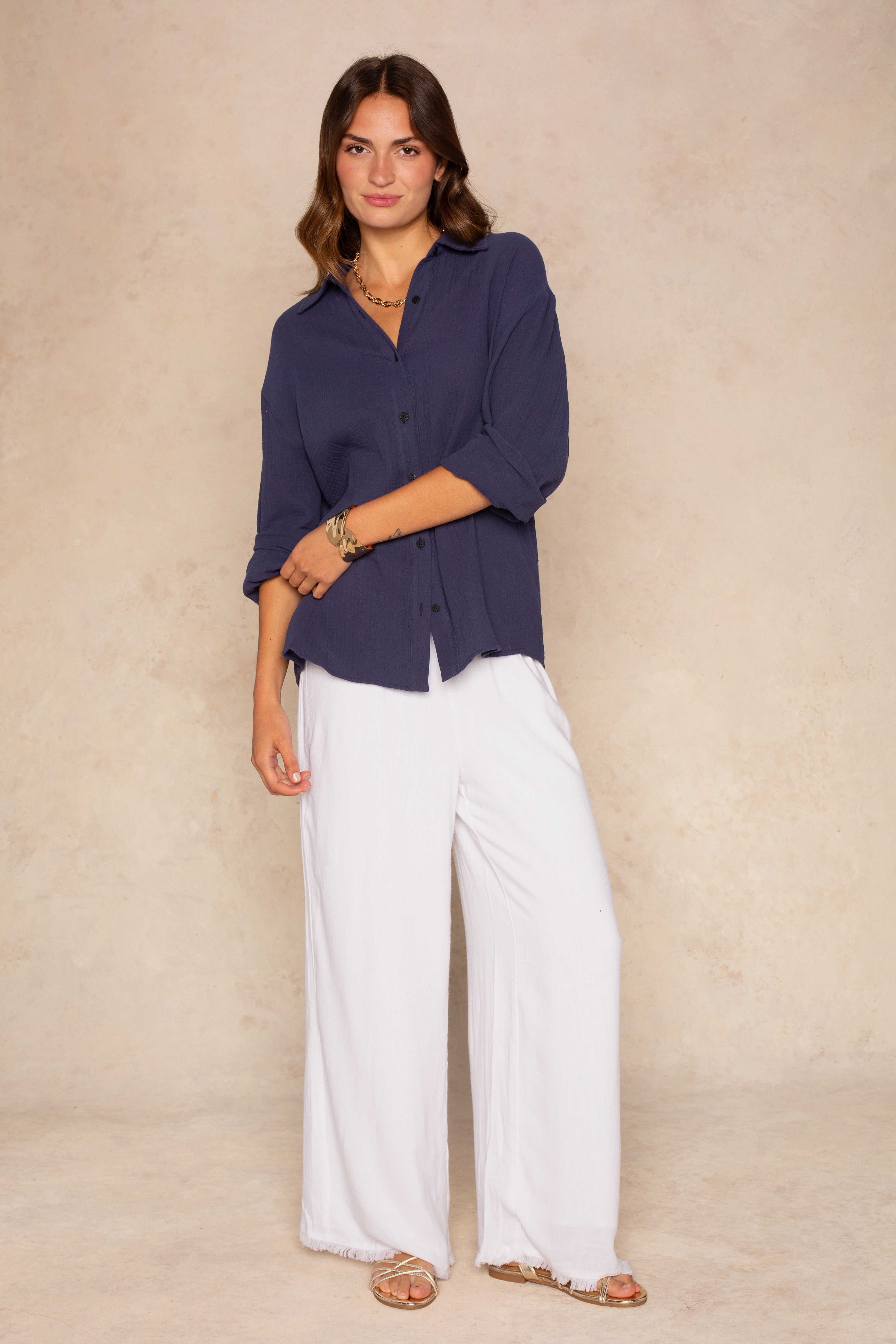 A woman with wavy brown hair, smiling slightly, stands against a beige background wearing the Isla Gia Gauze Button Down Shirt Coverup in navy, styled with white wide-leg pants, sandals, and gold jewelry.