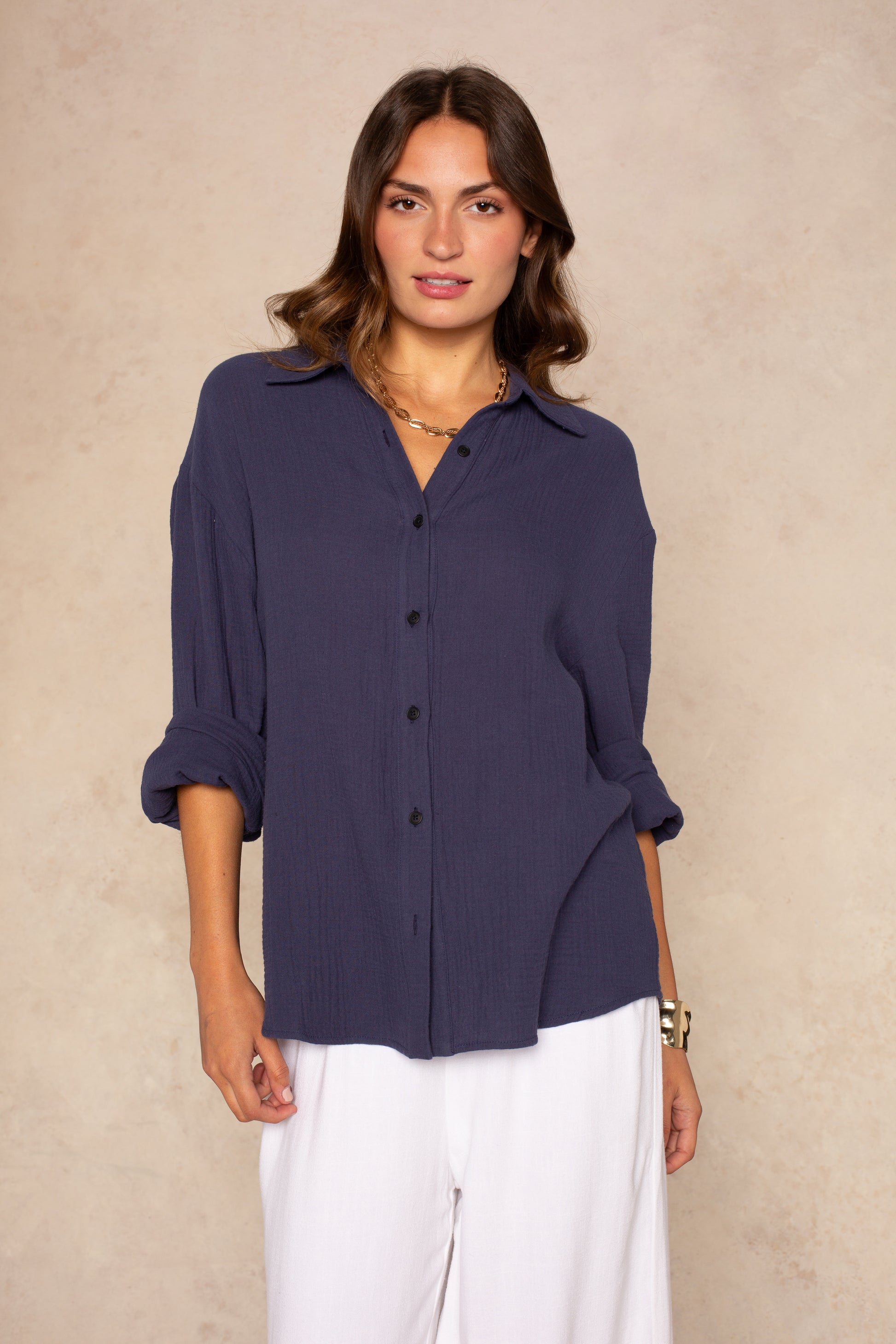A woman with wavy brown hair wears the Isla Gia Gauze Button Down Shirt Coverup in navy and white pants, standing against a beige textured background with a relaxed pose and neutral expression.