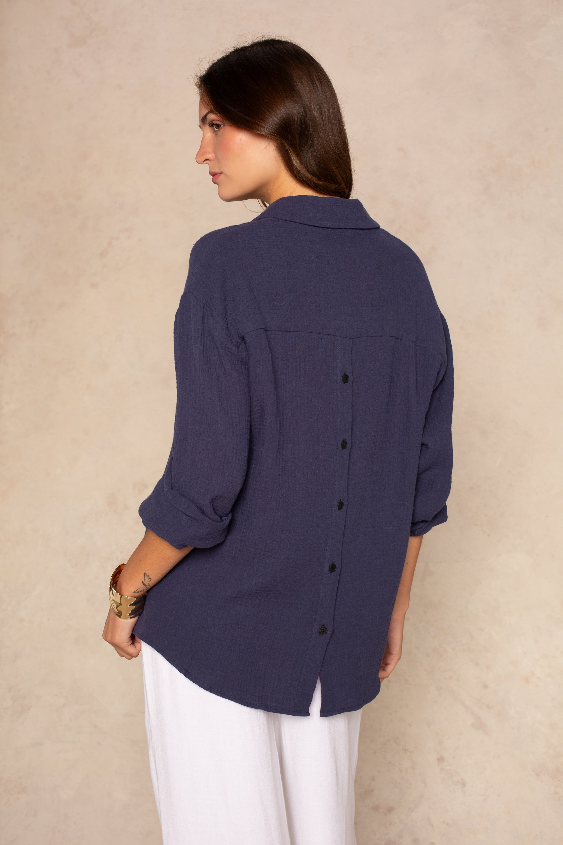 A woman with long brown hair, facing away, wears the Isla Gia Gauze Button Down Shirt Coverup in navy with black back buttons and white pants. The blouse is oversized and lightweight. Background is light beige.
