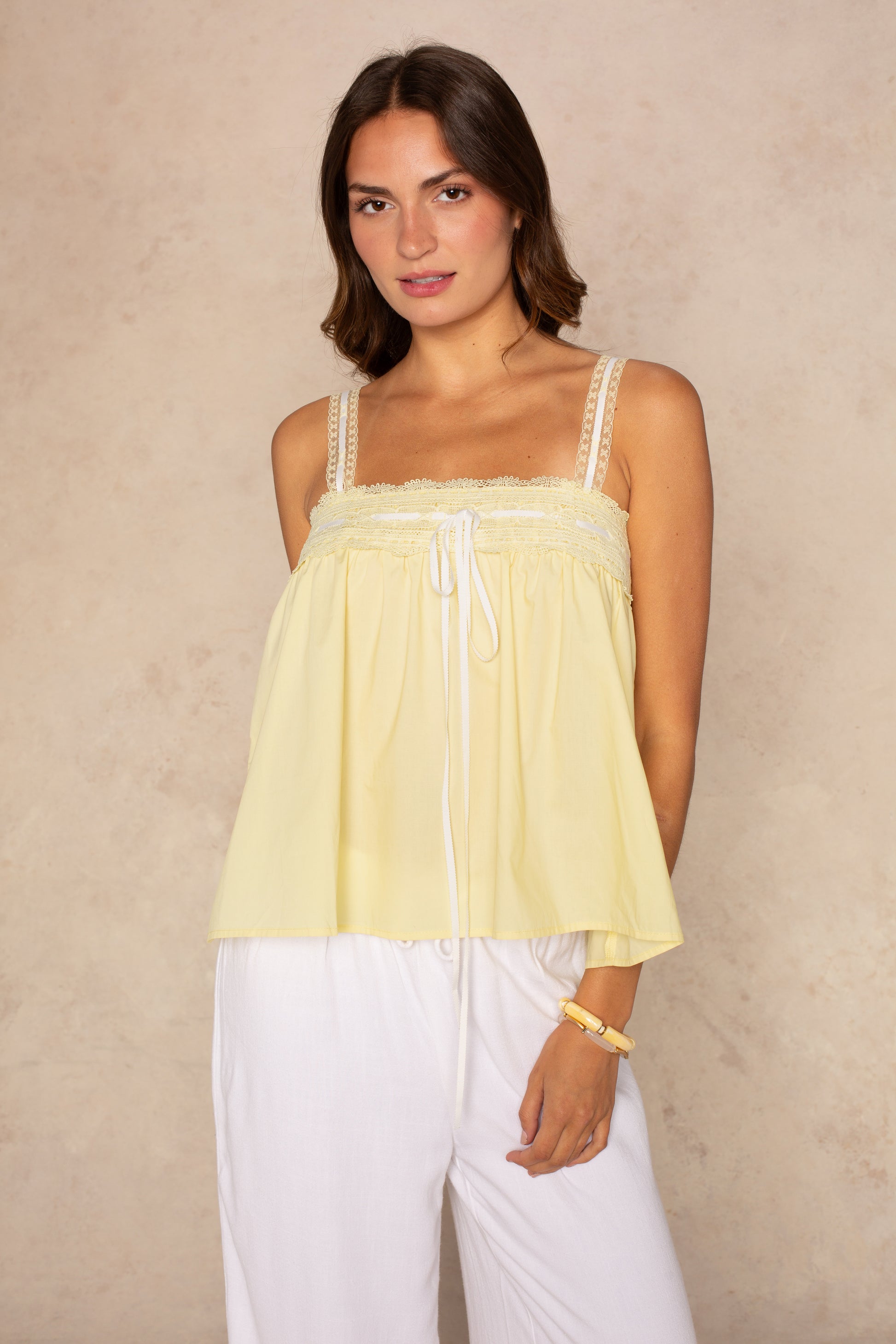 A woman with shoulder-length brown hair wears the Isla Mary Lace Trim Contrast Tie Tank in yellow, paired with loose white pants, standing against a light beige textured background.