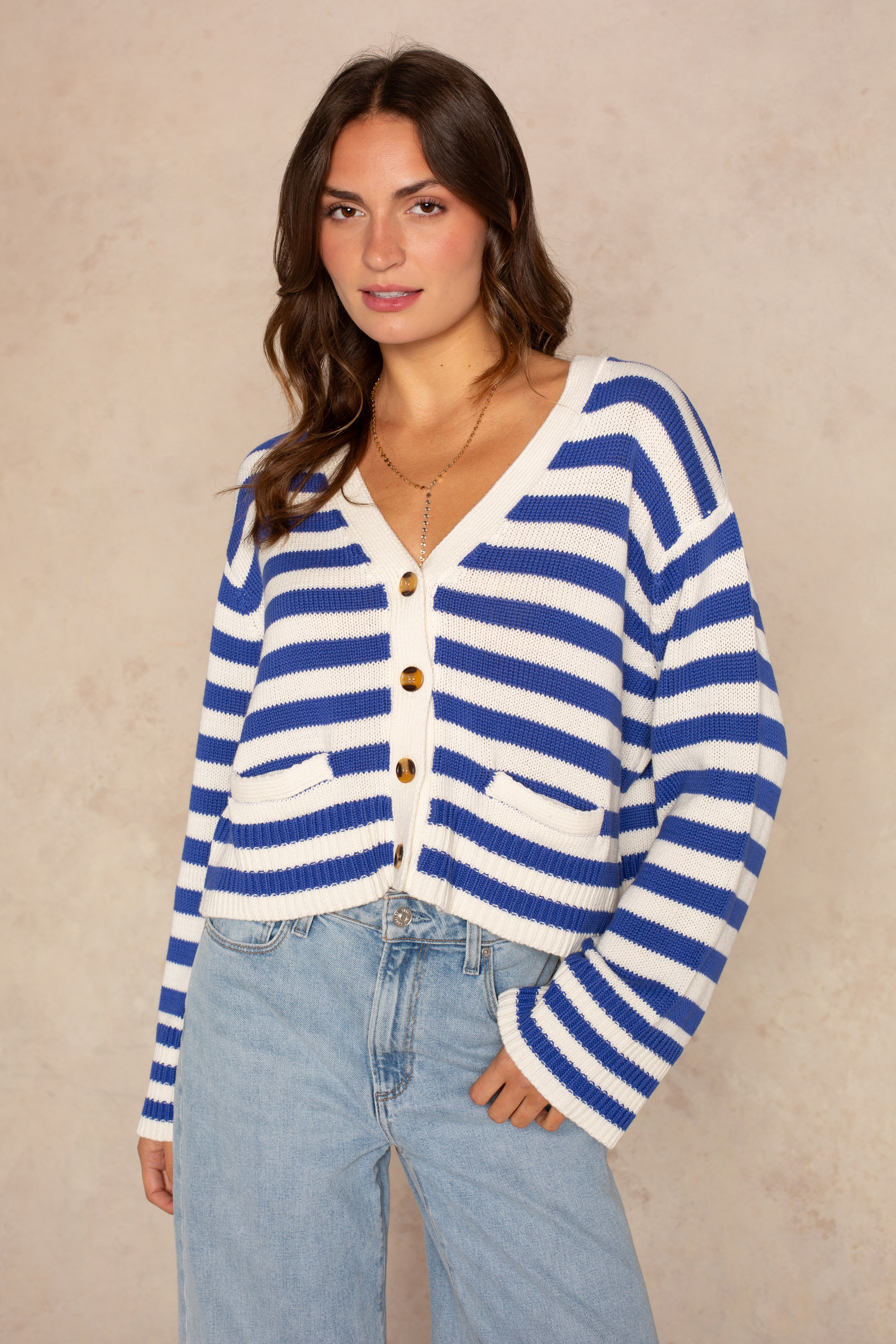 A woman wears the Isla Heather Stripe Crochet Cardigan in Blue Multi with front pockets and light blue jeans, standing against a beige textured background and smiling slightly at the camera.