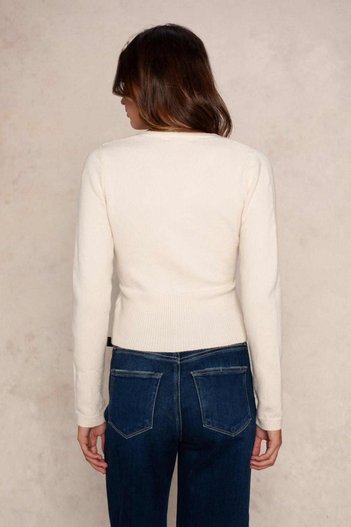 Woman wearing a cream ribbed wrap top with long sleeves paired with dark blue jeans, shown from behind.