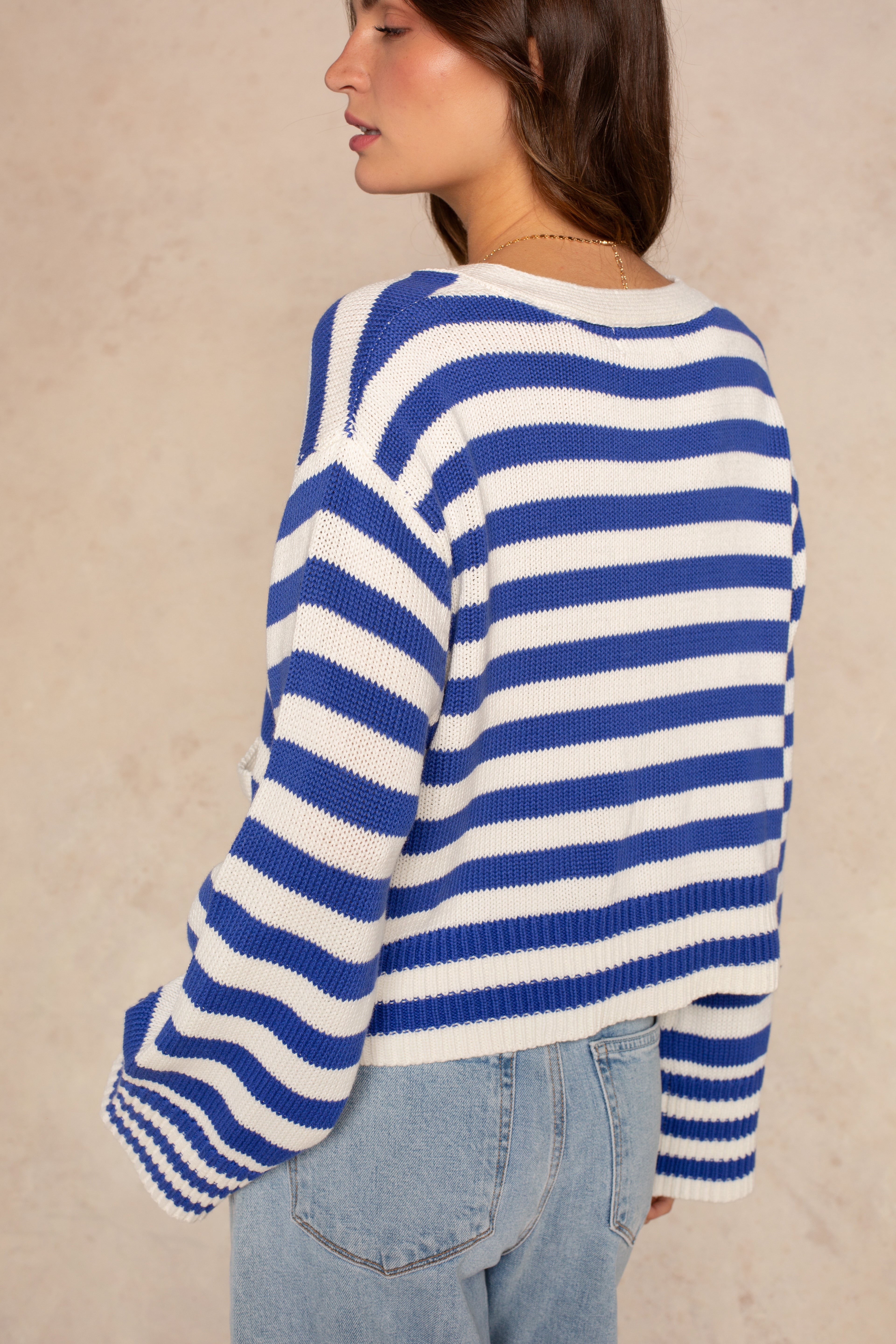 A woman with long brown hair, seen from the side and back, wears Isla’s Heather Stripe Crochet Cardigan in blue and white stripes with loose sleeves, paired with light blue jeans against a neutral beige background.