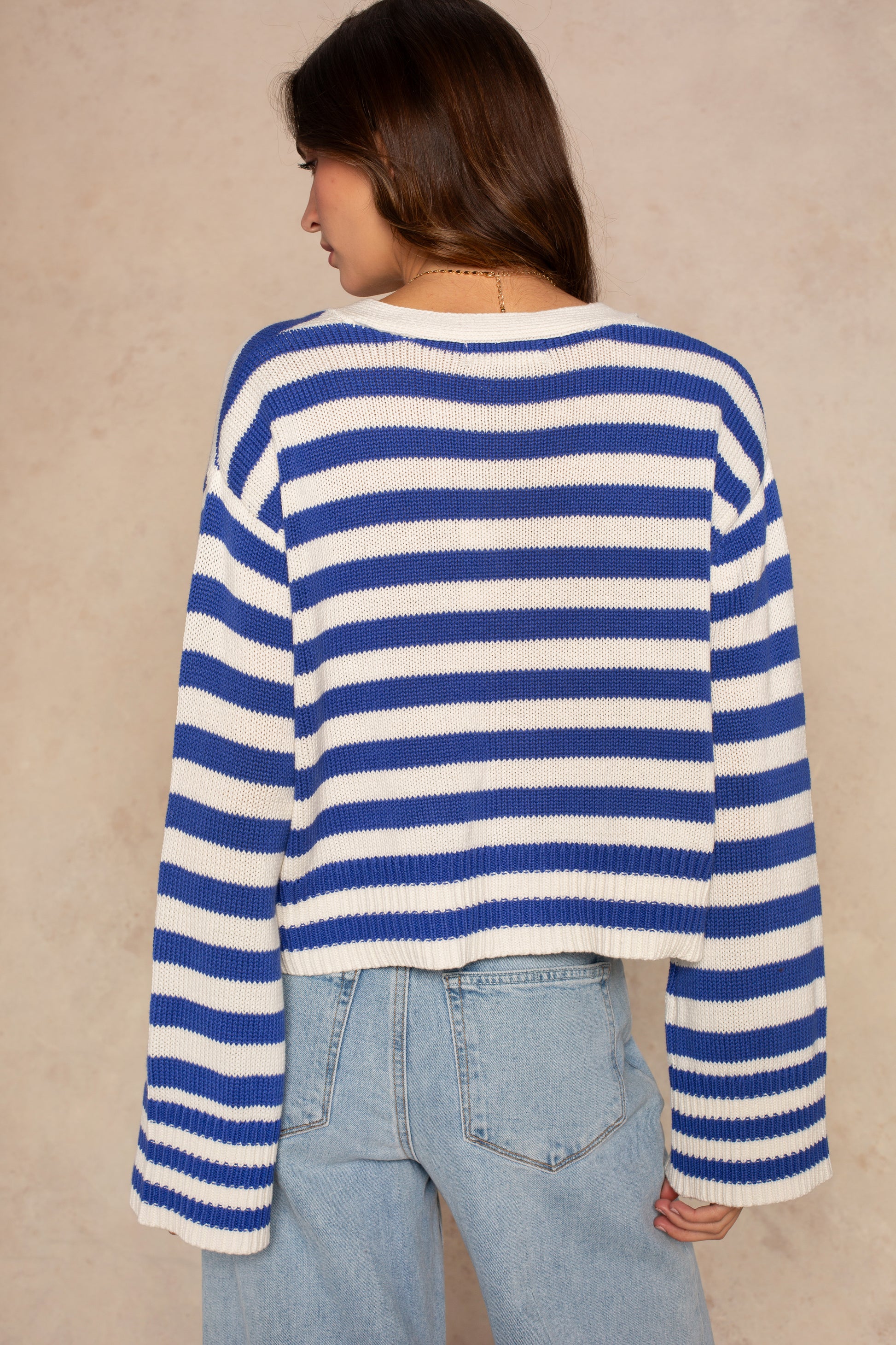 A woman with long brown hair, facing away from the camera, wears the Isla Heather Stripe Crochet Cardigan and light blue high-waisted jeans against a beige background.