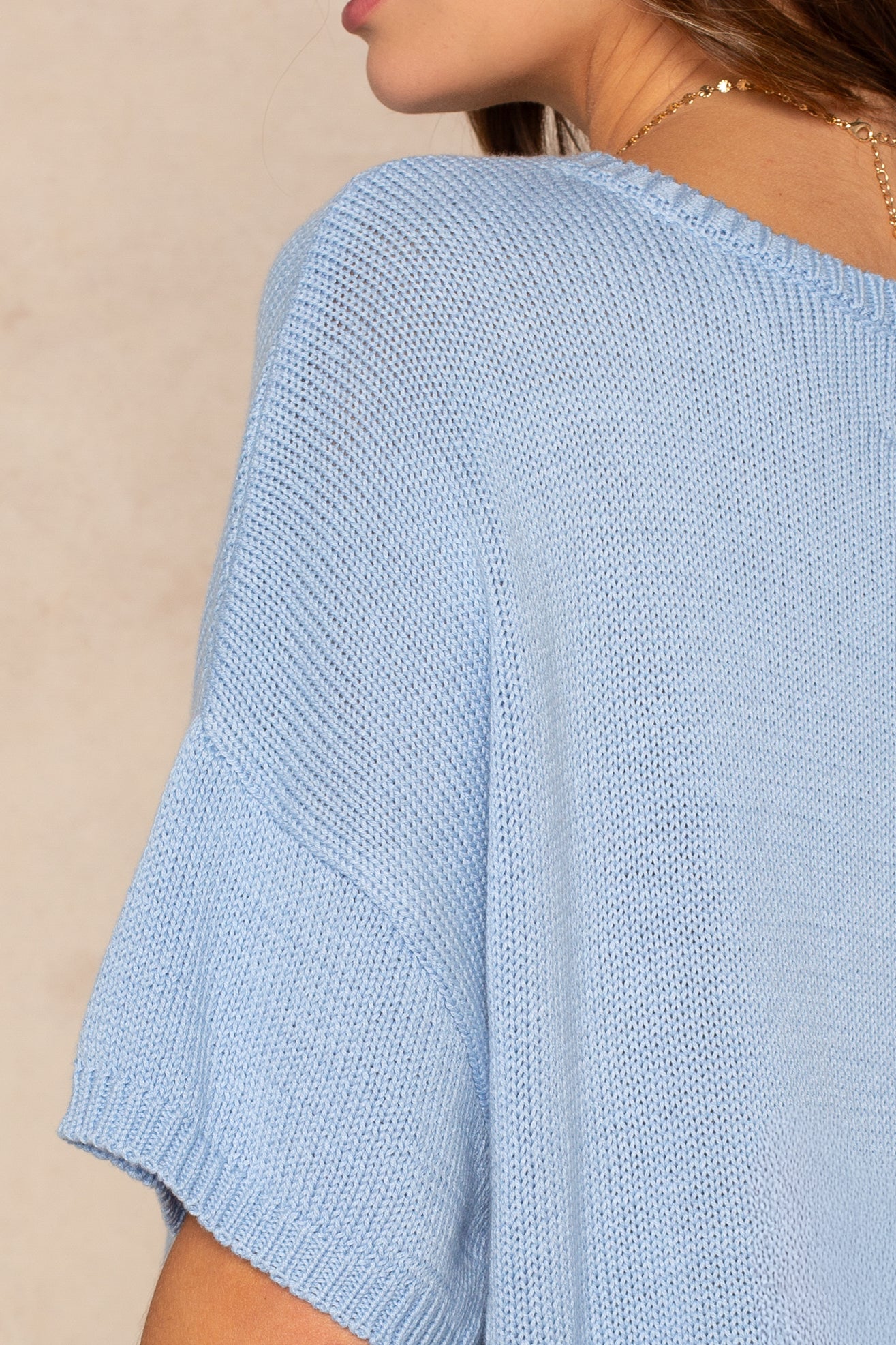 A close-up of a person wearing the Isla Lola Square Body, a light blue oversized knit top with a casual crew neck, styled with a gold chain necklace and photographed from behind against a neutral background.