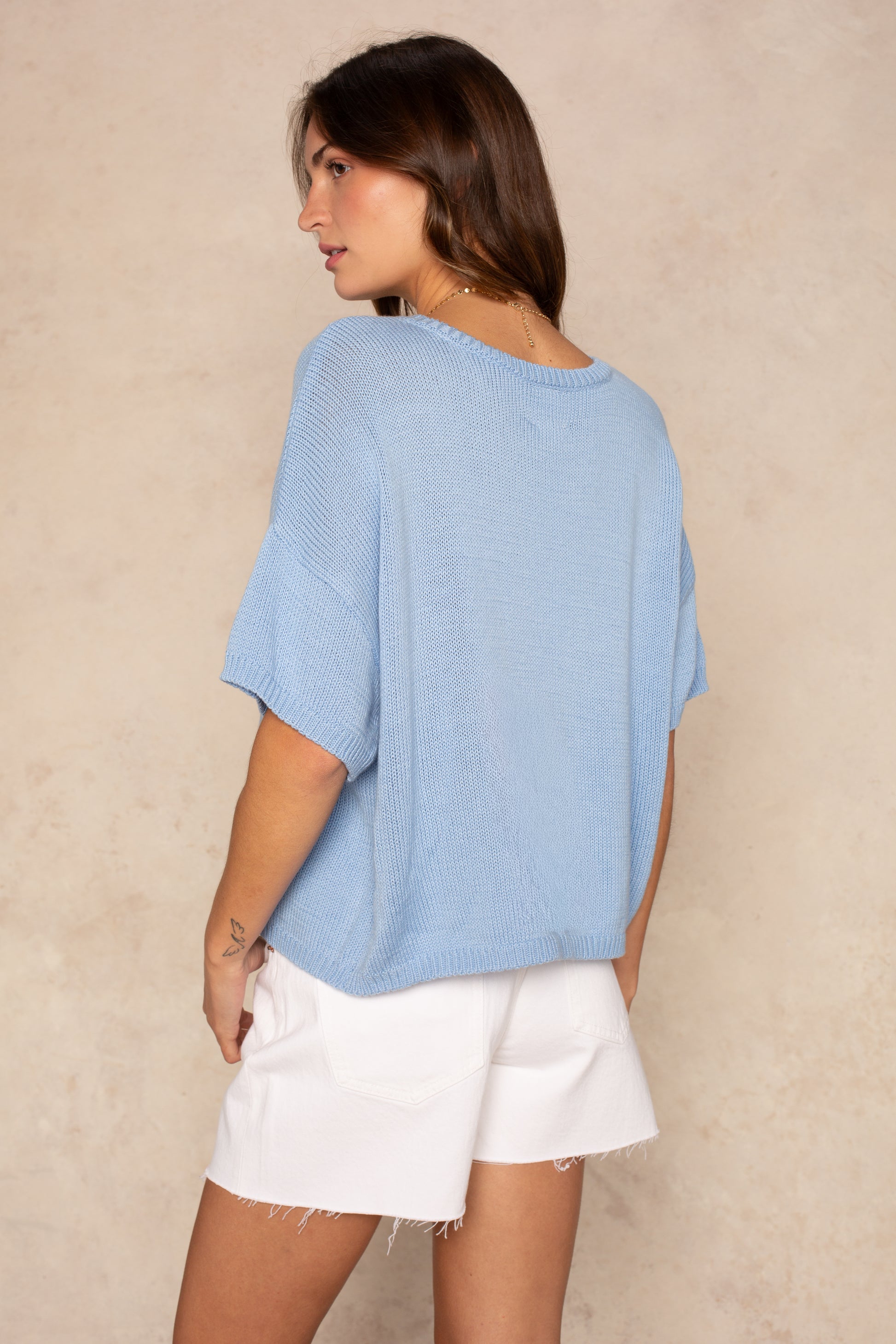 A woman with long brown hair models the Isla Lola Square Body, an oversized light blue knit sweater, paired with white frayed shorts as she stands sideways and looks over her shoulder against a beige textured background.