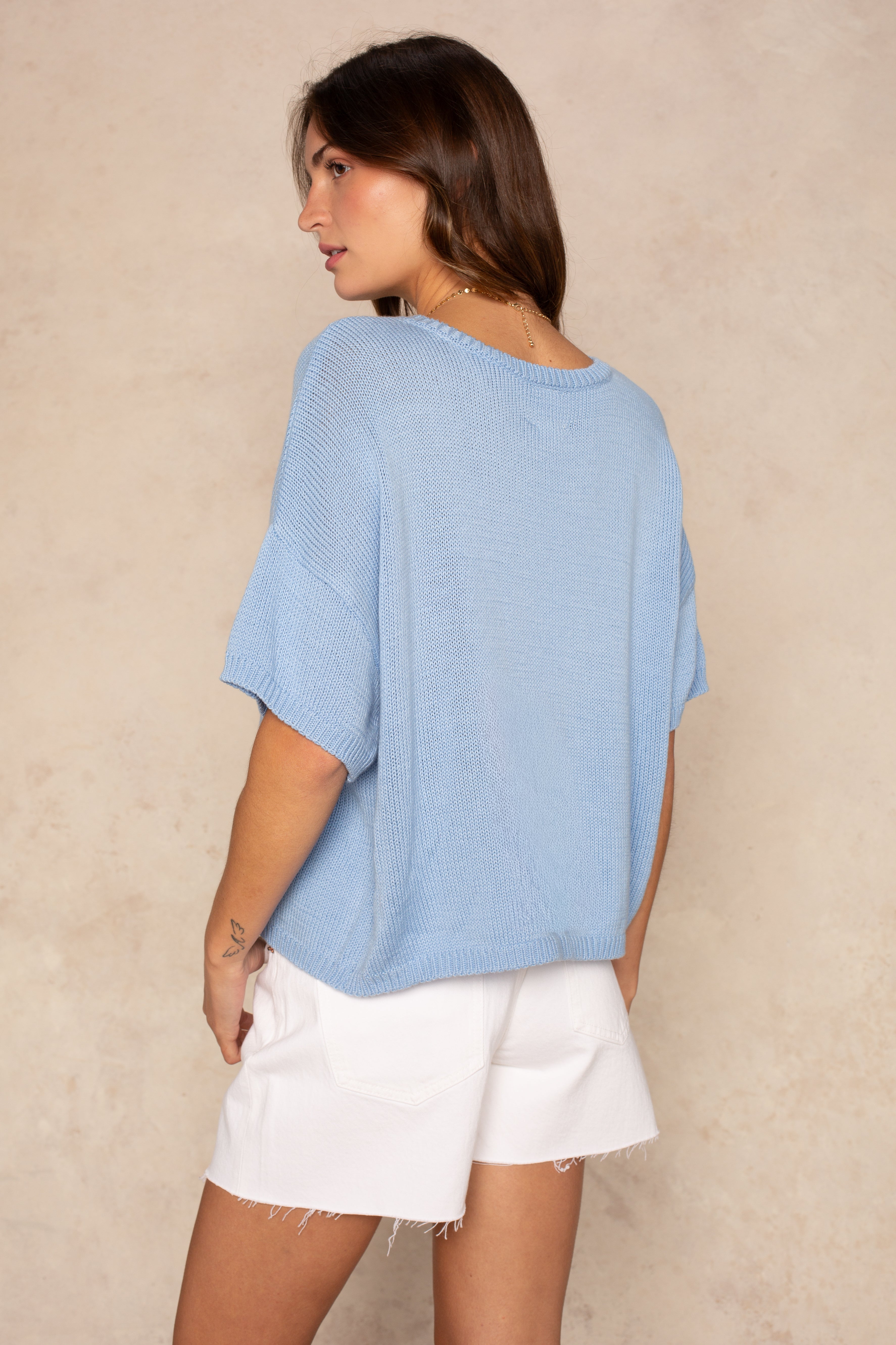 A woman with long brown hair models the Isla Lola Square Body, an oversized light blue knit sweater, paired with white frayed shorts as she stands sideways and looks over her shoulder against a beige textured background.