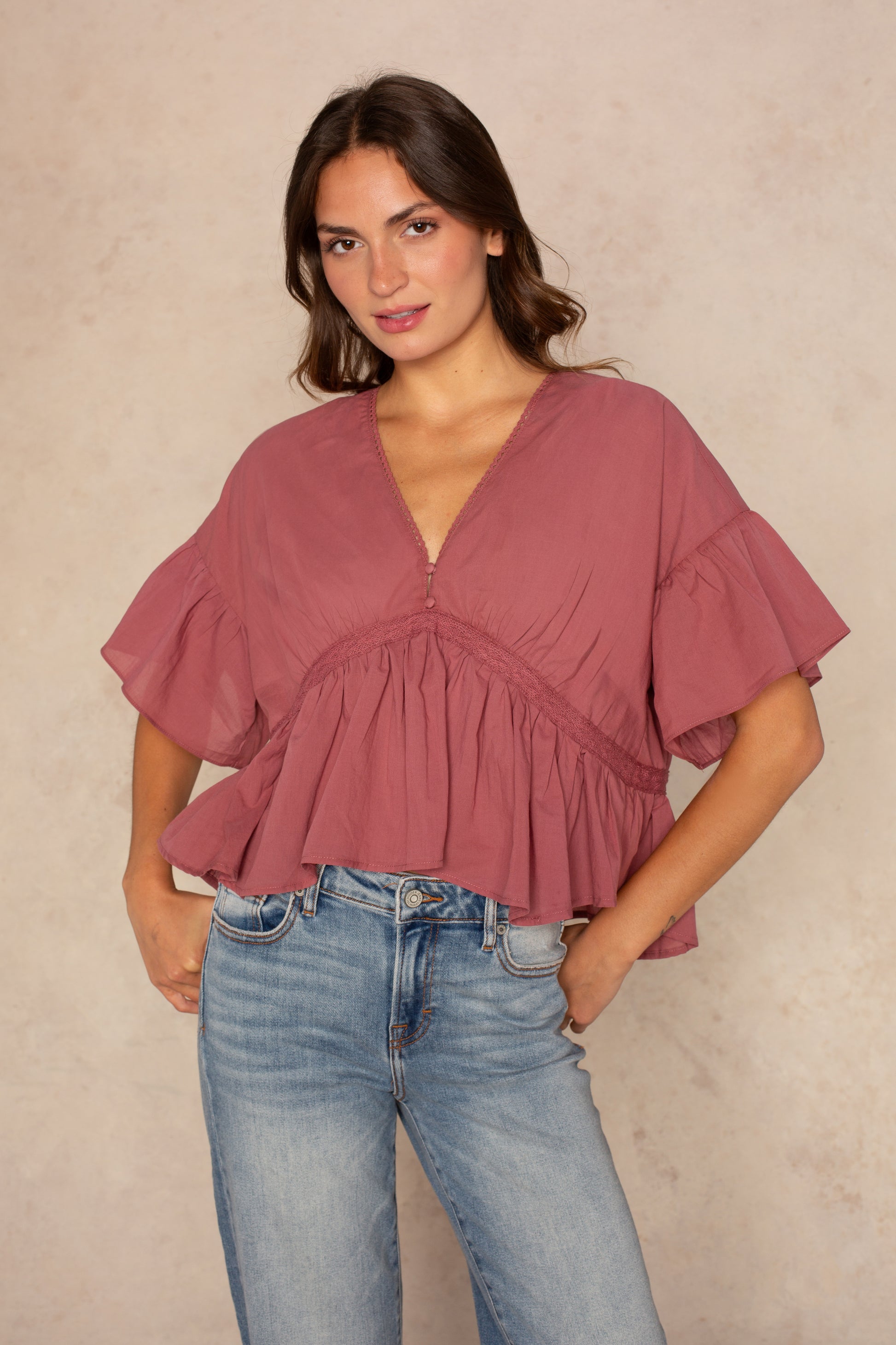 A woman wearing the Isla Hazel Blouse in Red Multi and blue jeans stands against a light beige background, looking at the camera with a relaxed expression.