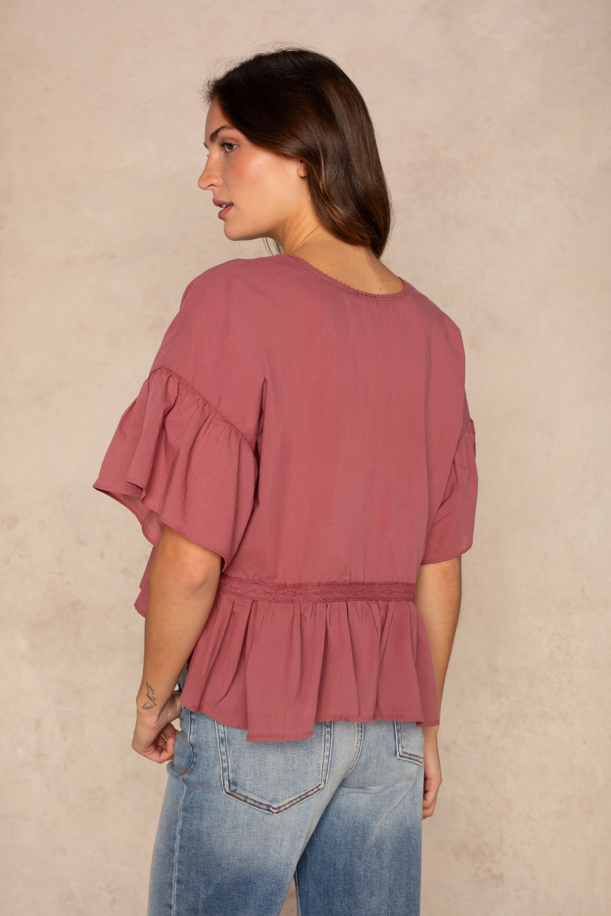 A woman with long brown hair stands sideways against a beige background, wearing Isla's Hazel Blouse in dusty rose with ruffle sleeves and hem, paired with light blue jeans.