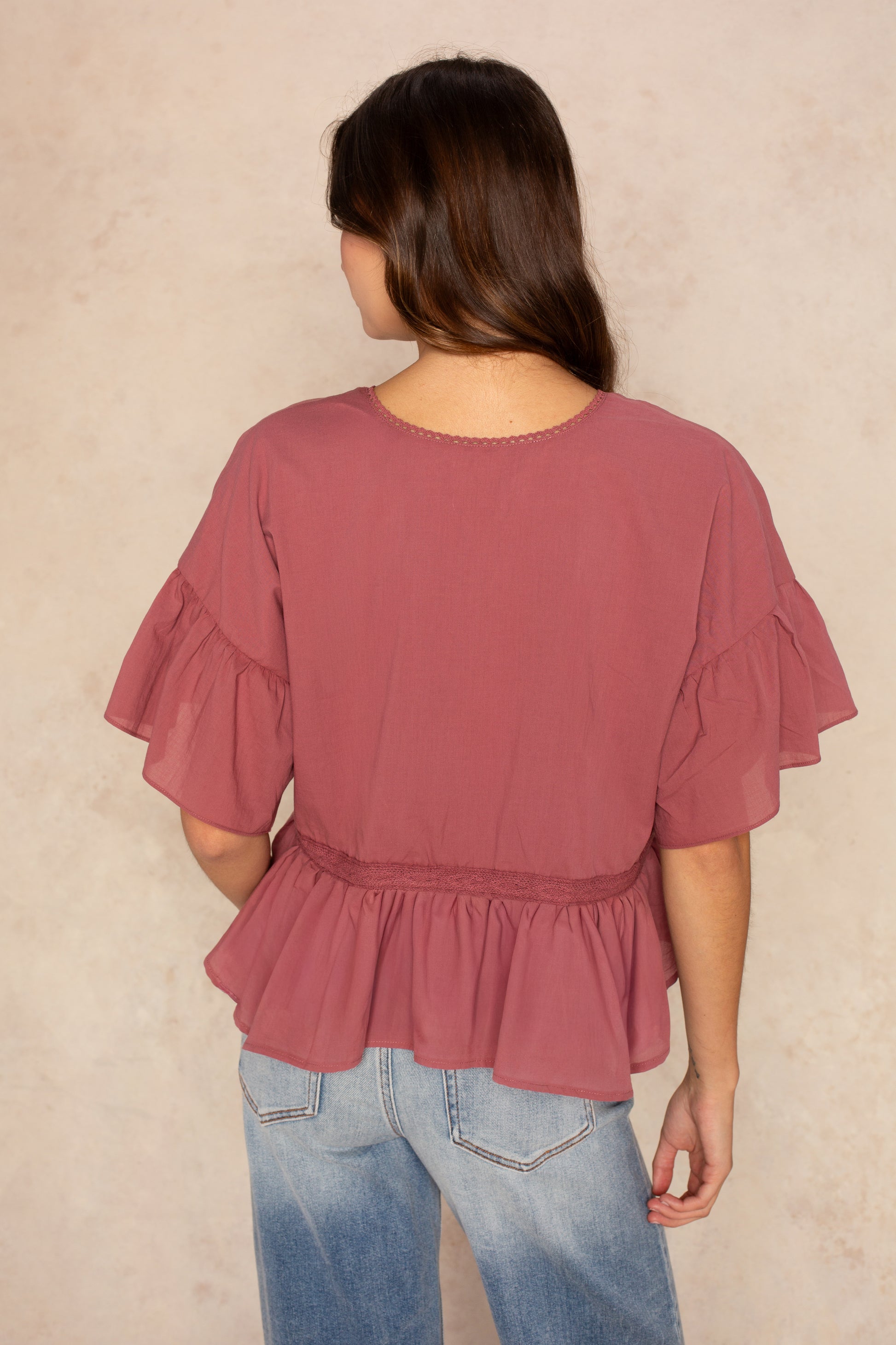 A woman with long brown hair, seen from behind, wears the Isla Hazel Blouse in dusty rose with ruffle sleeves and light blue jeans, standing against a light beige background.